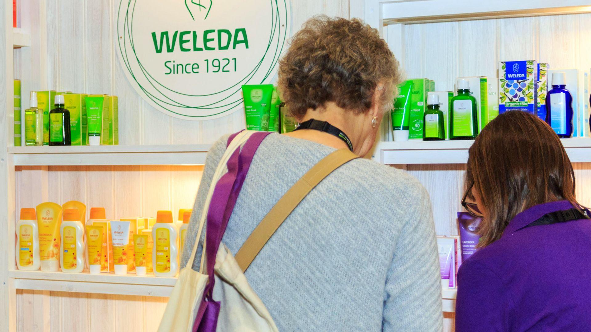 Two people stand in front of a display shelf featuring various Weleda products arranged by colour: green, yellow, pink, and blue. A circular logo in the centre reads "WELEDA Since 1921" with an abstract plant design above it. The person on the left wears a light grey sweater and carries a tote bag with purple straps; the person on the right wears a purple shirt. They are both facing the shelves so only their backs are visible.
