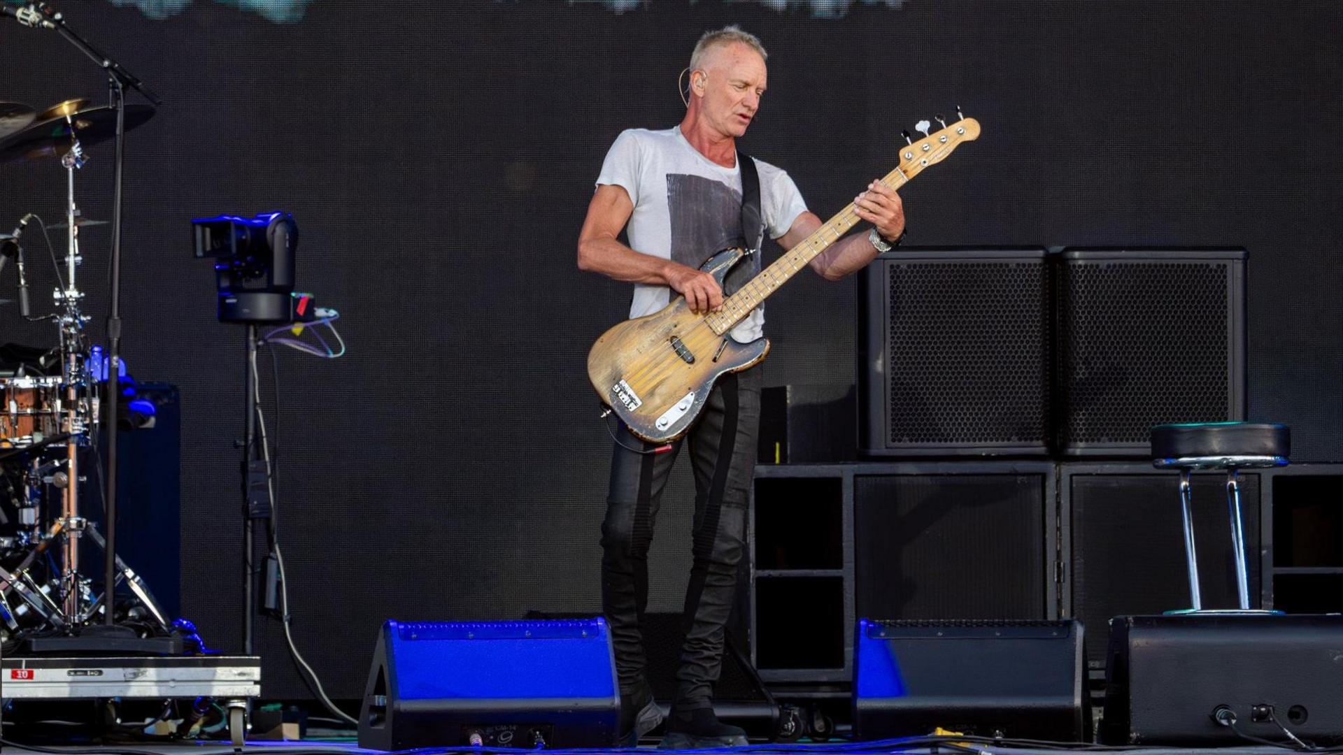 Sting performing at the Isle of Wight Festival on Friday. He has short, cropped grey hair and wears a white t shirt and tight dark trousers. He's playing a bass guitar.