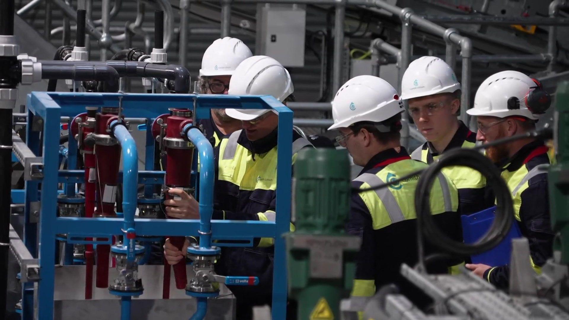 A number of people stood together working on an item in a warehouse. They are all wearing hard hats, glasses and a hi-vis.