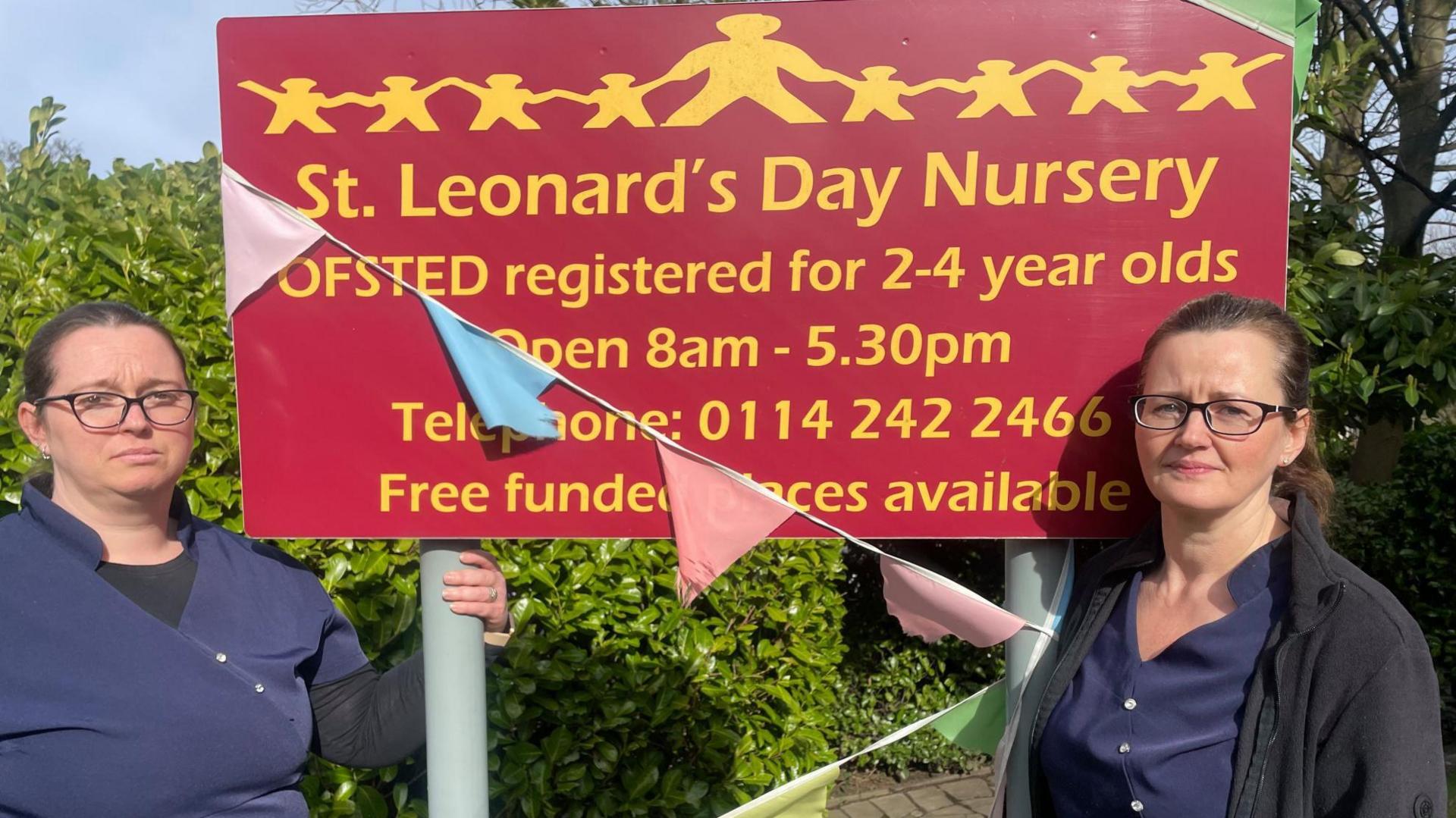 Two women stand either side of a crimson sign wearing blue uniforms. They look upset.