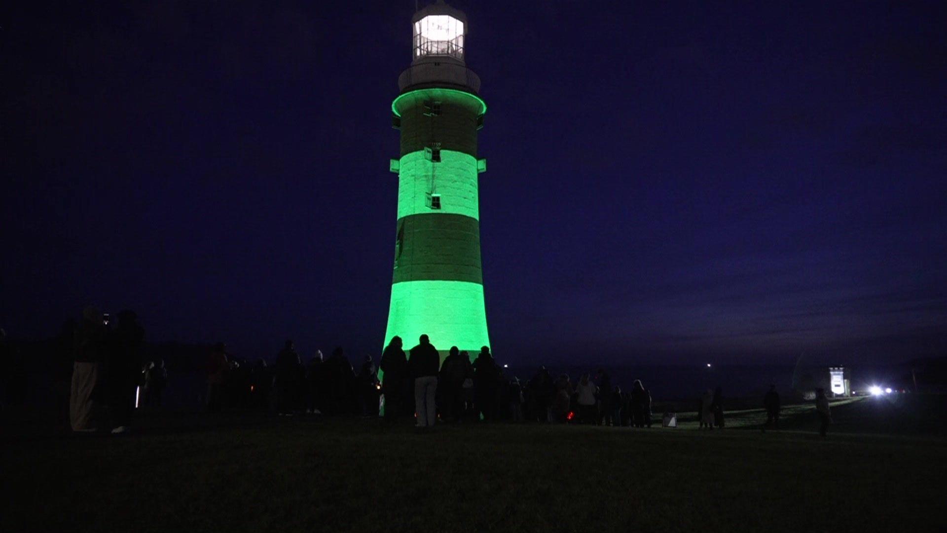 A lighthouse is lit in a green light at dark with a number of people gathered in front of it.