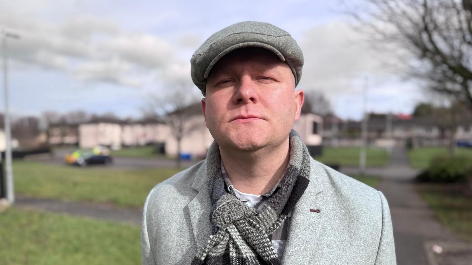 A man wearing a grey coat, scarf, and cap standing outside with some trees and houses in the background. He is looking at the camera with a serious expression