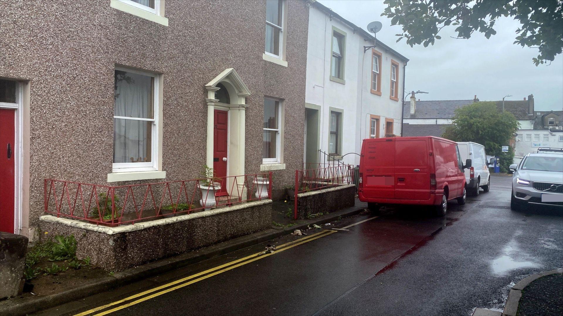 A picture of Adam Carruthers childhood home which is a terraced house with pebbledash and a central red door. There is no pavement so from the door there is a small, walled space which steps straight on to the road. Two vans are parked outside and a silver Volvo is driving towards the camera through a narrow gap.