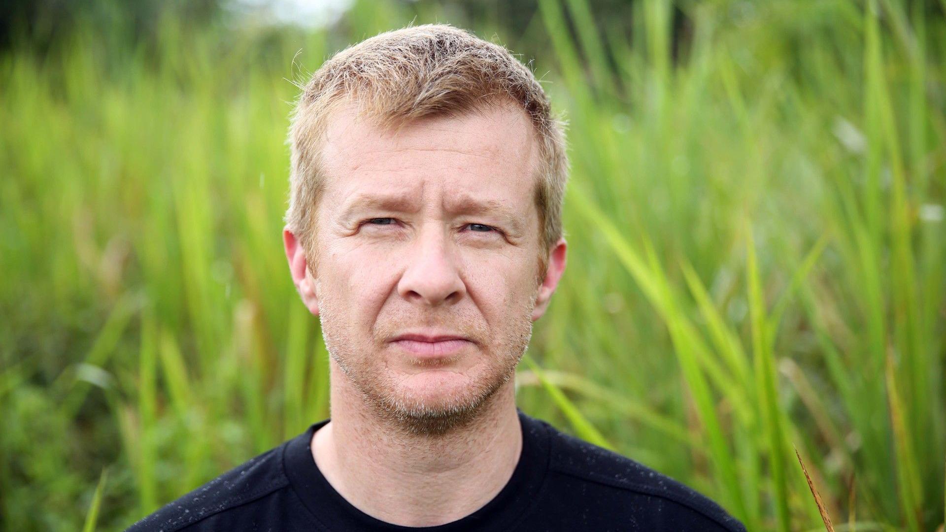 A man with stubble and cropped blonde hair stares seriously into the camera 