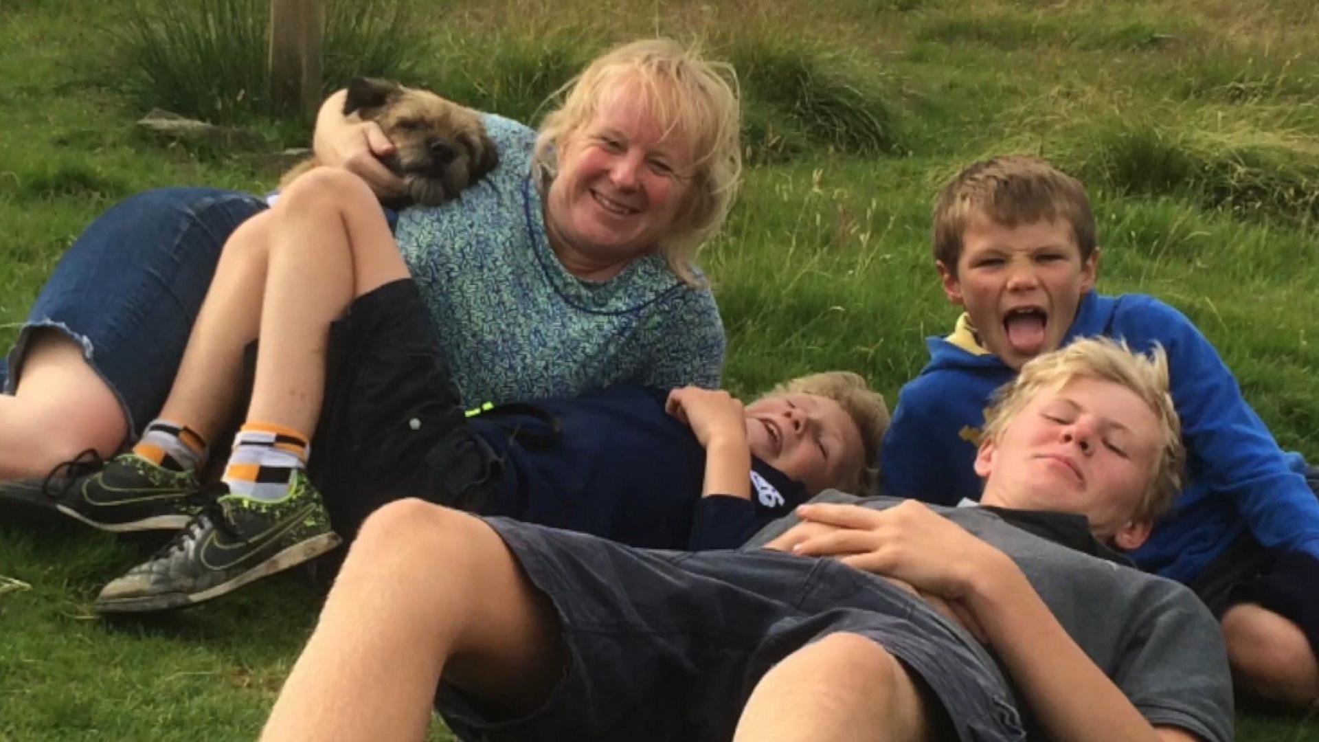 A boy with blonde hair, lying on the ground, leans back against a dark-haired boy with another blonde haired boy lying next to them and a woman with blonde hair cradles a small brown dog. All are smiling at the camera