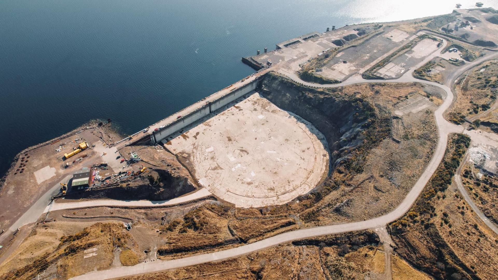 Aerial view of Kishorn Port showing its dry dock. The dry dock is a bowl shape cut into the rocky coastline. The bottom of the dry dock is a flat expanse of concrete. There is a road above the dock.