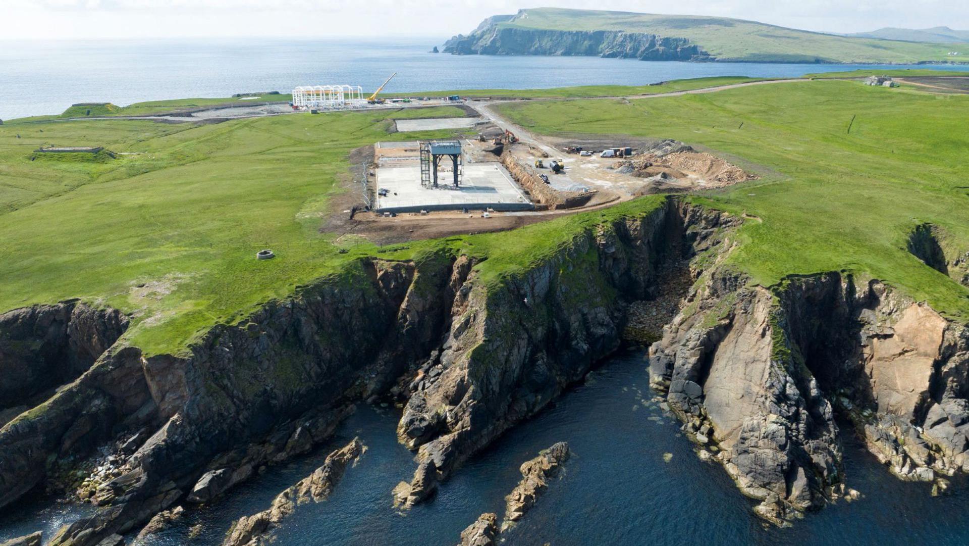 An aerial image of SaxaVord spaceport under construction on the island of Unst in Shetland. There is a launch pad with a launch tower on a flat area of land above sheer cliffs to the sea below.