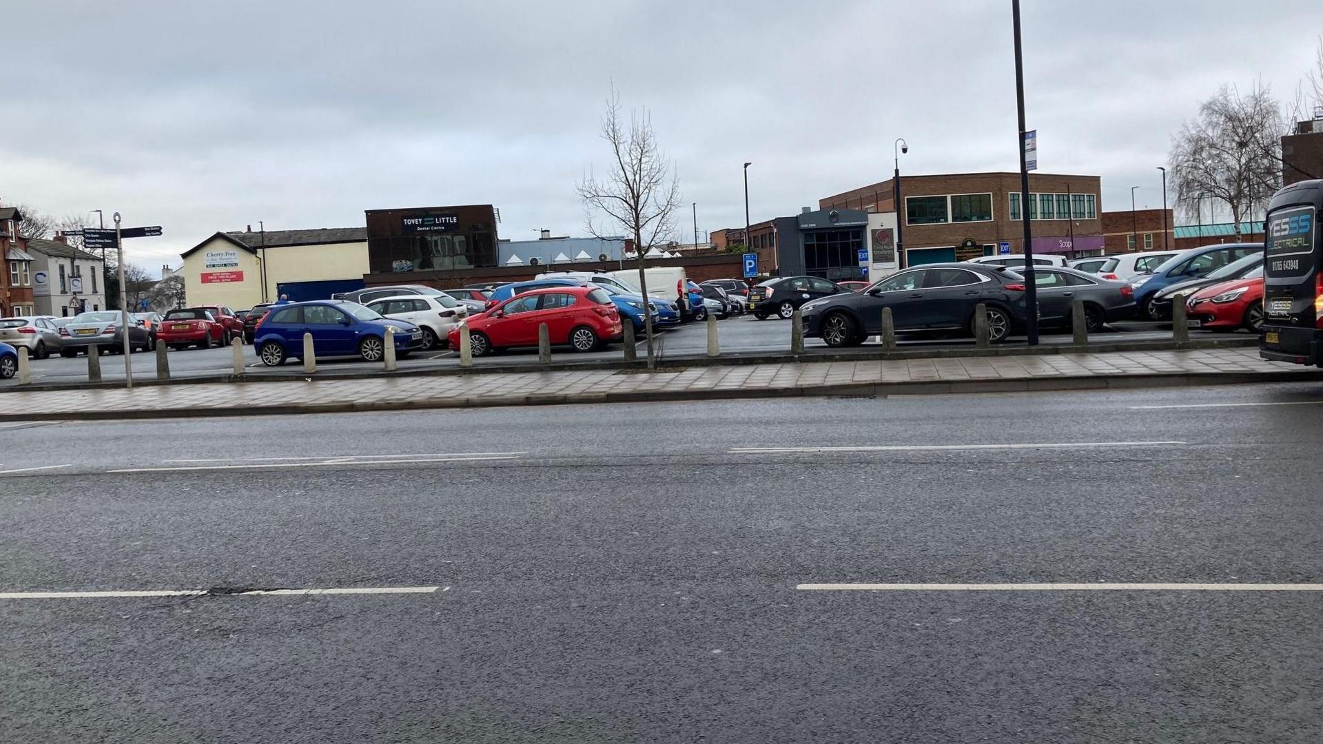 A public car park full of cars, pictured from the other side of the road on a cloudy day.