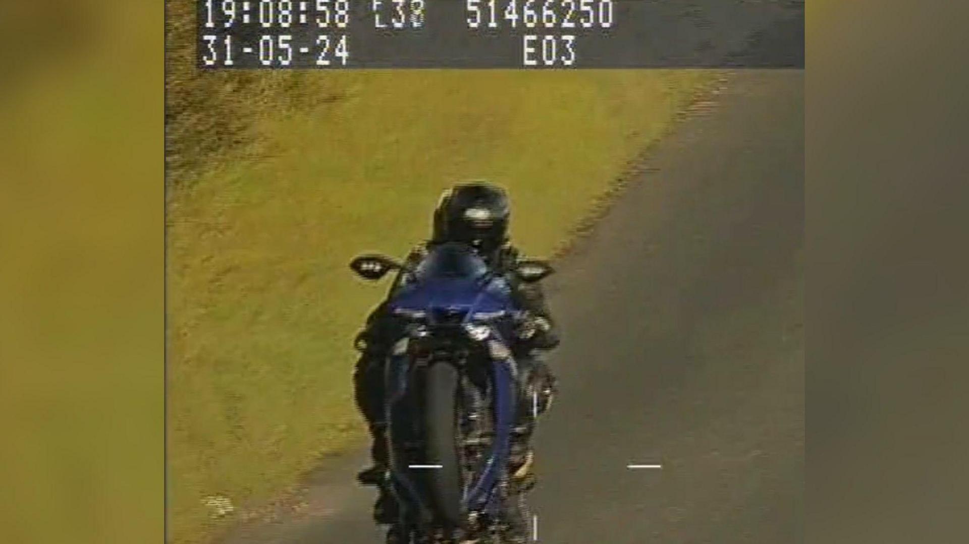 A picture of a person riding a black and blue motorbike on a road seen with the front wheel of the motorbike in the air. 