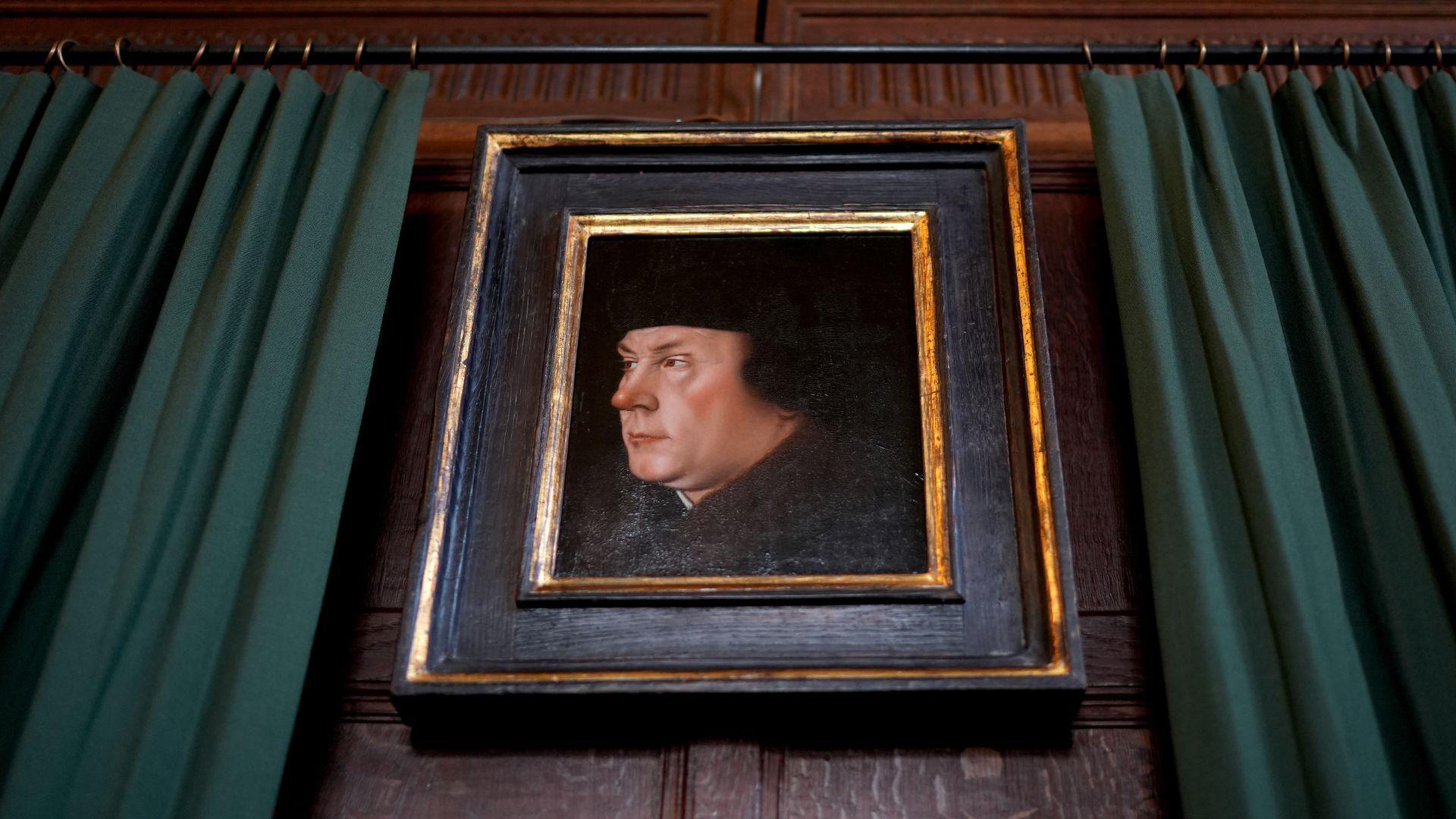 A framed portrait of Thomas Cromwell on a dark wooden wall. The frame is dark brown wood with gold trim. To the left and right of it are green curtains. A black curtain rail runs above the portrait