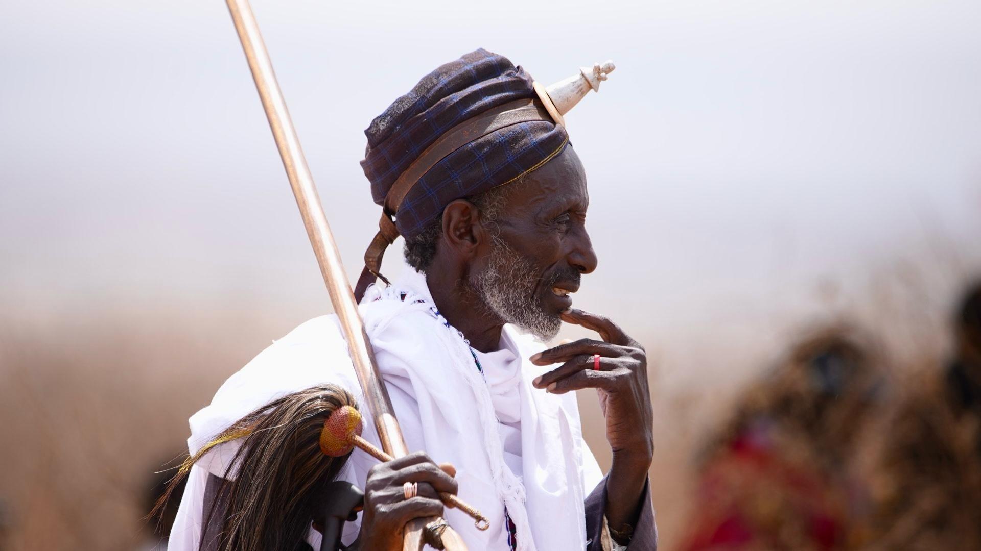 An elder wears a headdress and carries items that indicate his status.