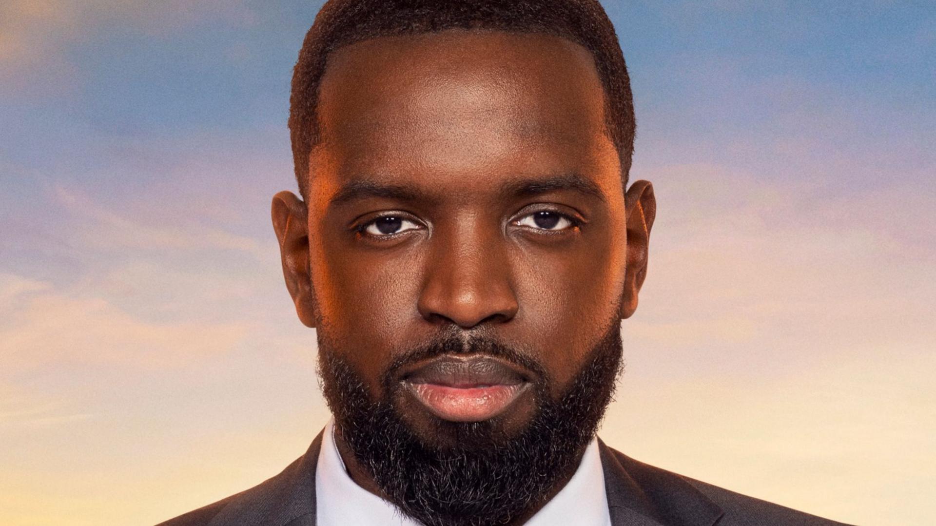 A publicity image where Frederick Afrifa has a tidy dark beard and dark short hair. He is wearing a dark grey suit with a dark red tie and crisp white shirt. Behind him is a sunset or sunrise and part of the London skyline.