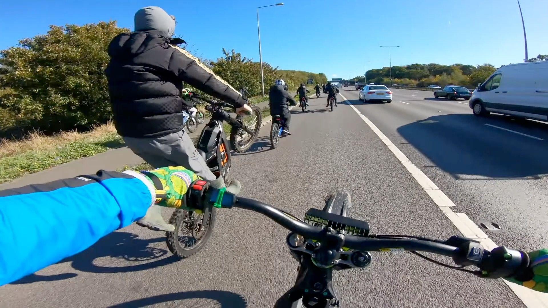 Ebike riders illegally drive down the M2 motorway, in a screen grab from a video. There is a group of riders on the hard shoulder, with traffic seen across three lanes to the right of them. The nearest rider is doing a wheelie.
