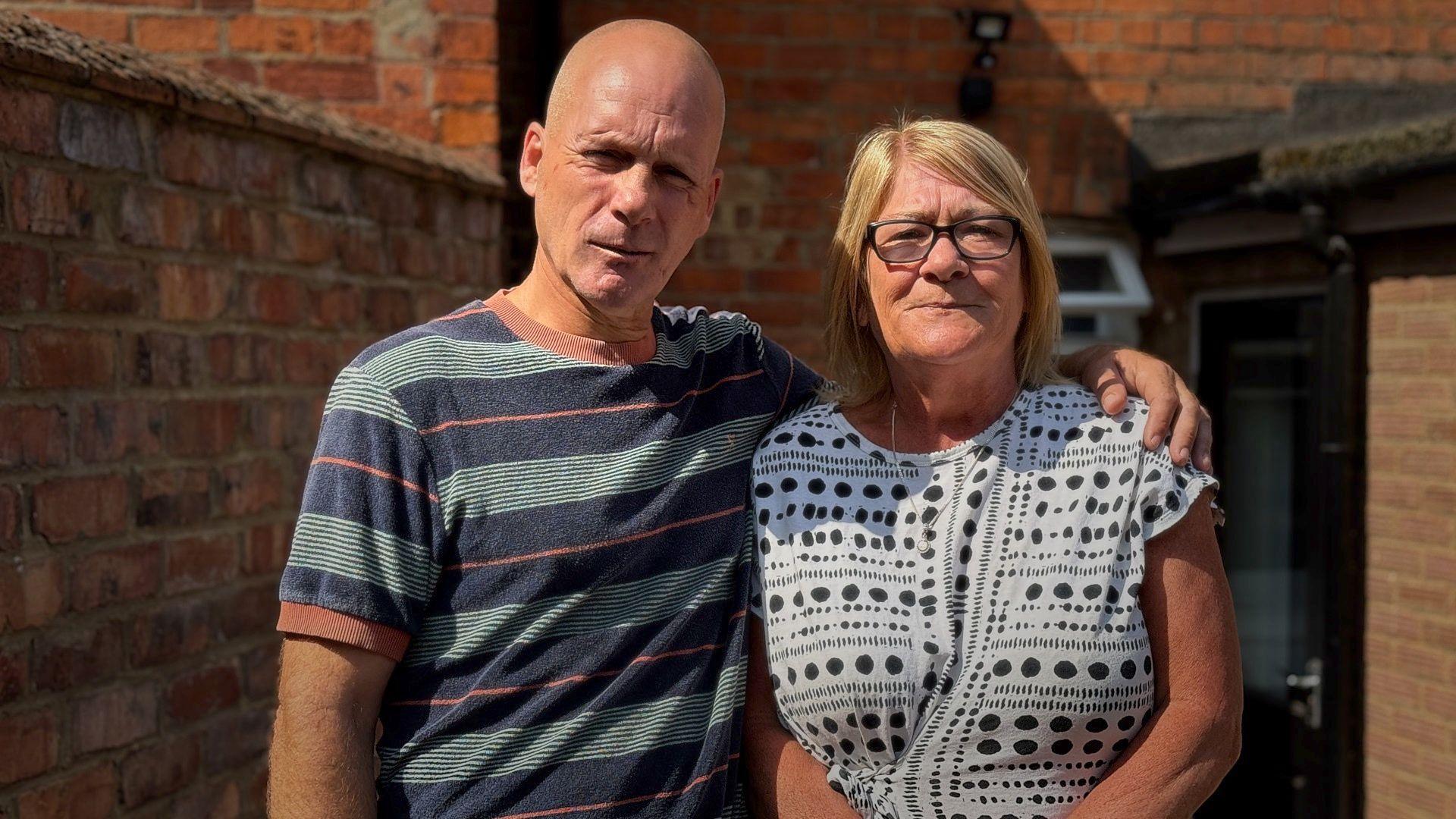 In front of a red brick wall, Lee, a bald-headed man in a blue and orange striped tee shirt, puts his arm around Theresa Kelly's shoulders, who wears a black and white dress and wears black-rimmed glasses.