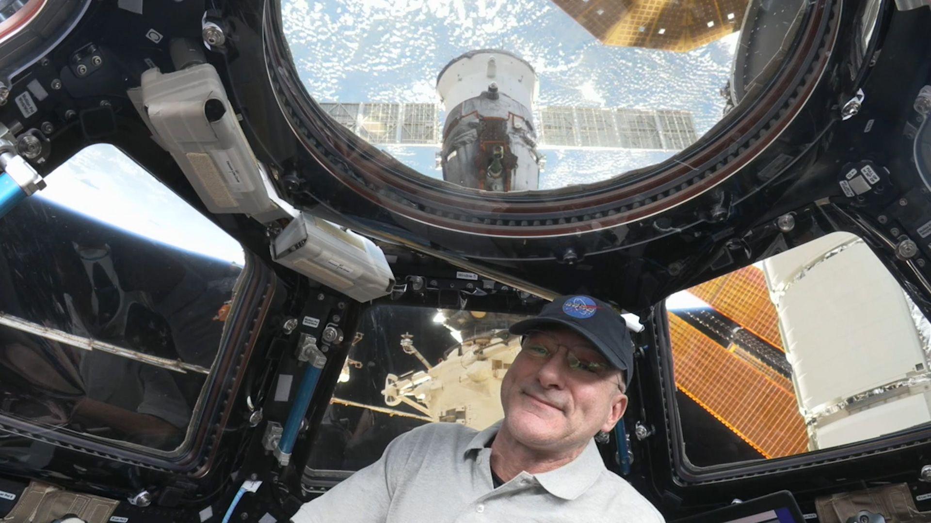 A man wearing a cap smiles on board a space station, where we can see the Earth through its windows