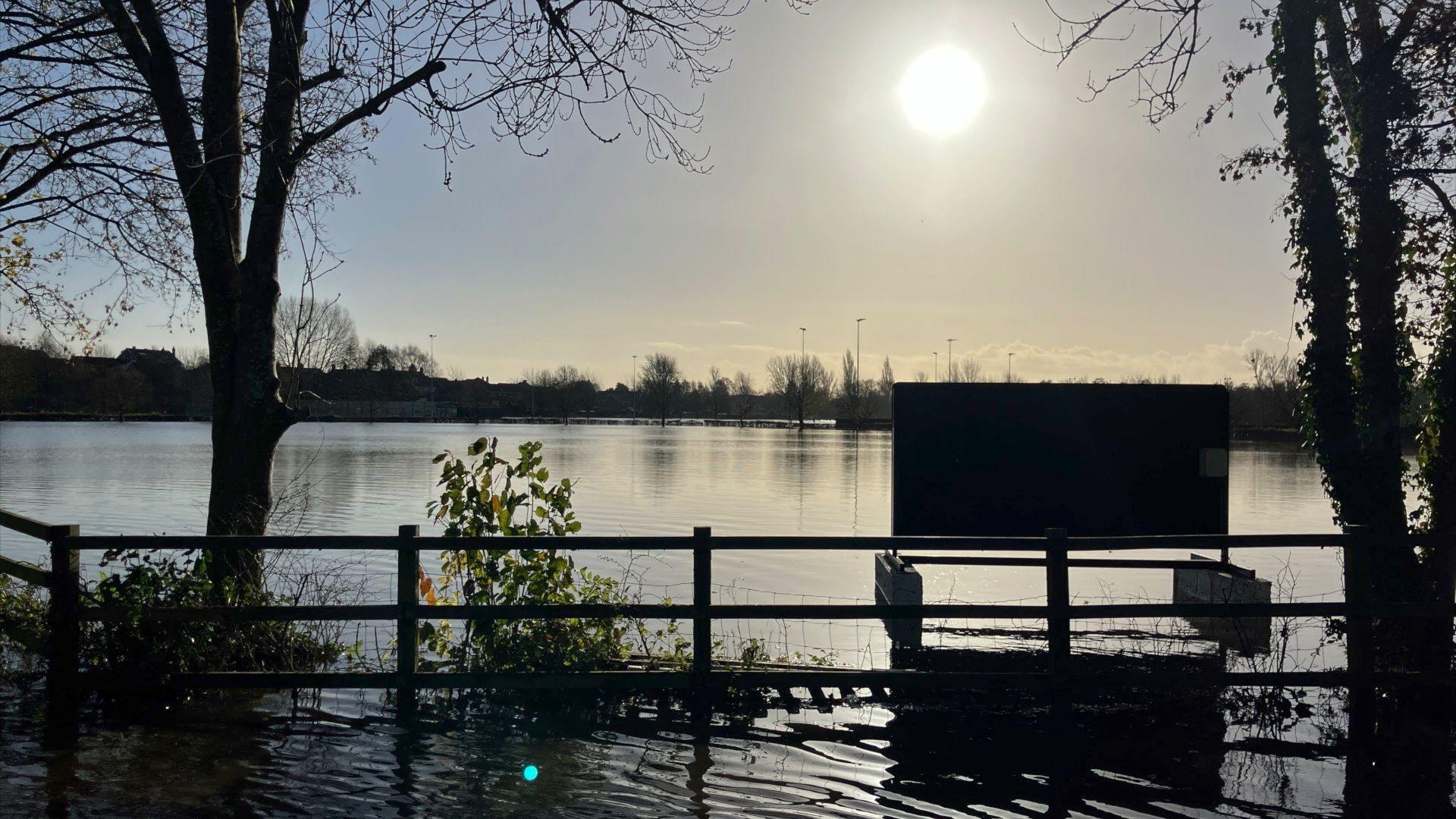 Flooding in Lydney showing fields submerged in water, with the sun low in the sky and trees silhouetted against the sky. A fence and a sign is also visible