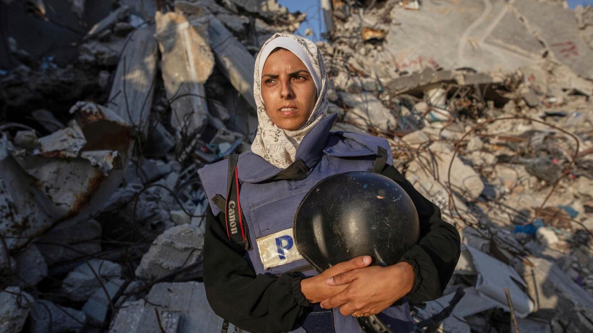 Journalist Marian Abu Dagga wears a blue press vest and holds a black safety helmet. She is standing in front of the rubble of a destroyed building, looking off to the distance with sun lighting her face.