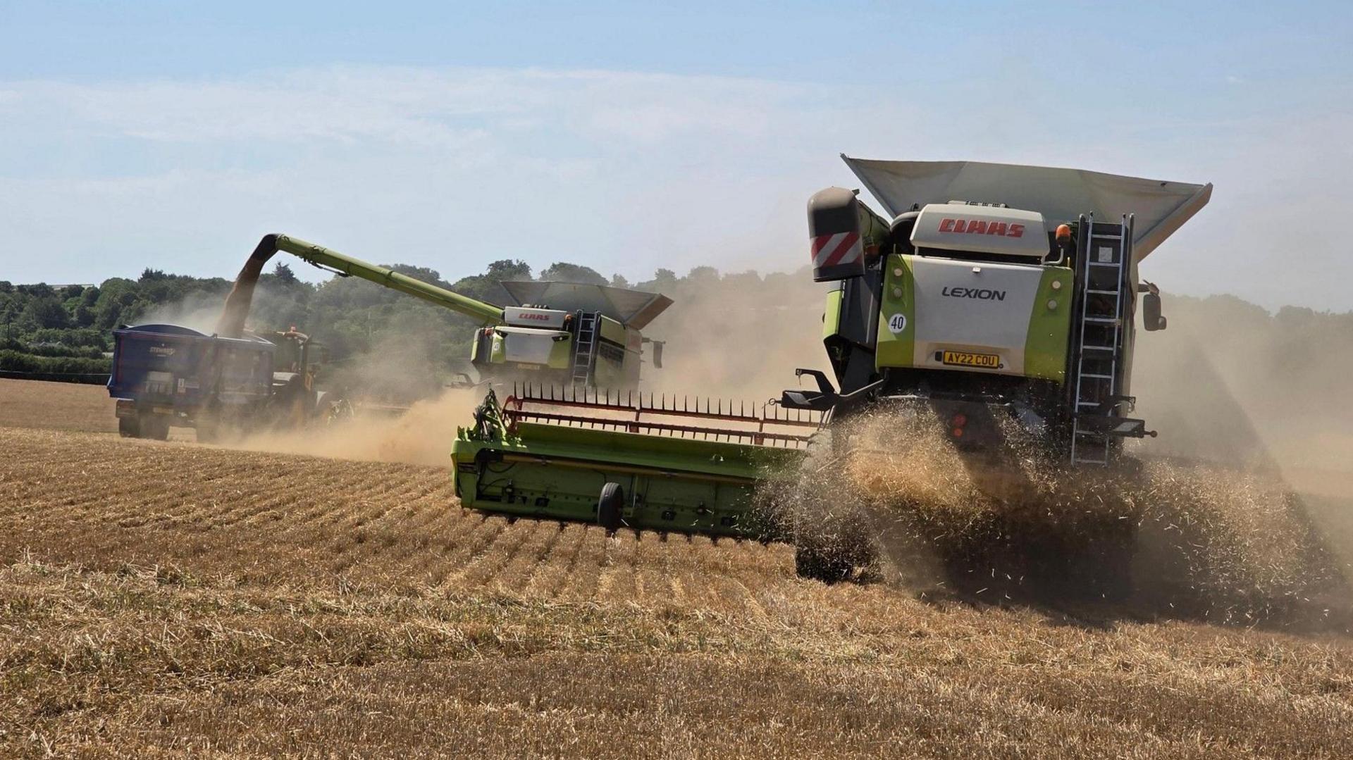 Farm machinery kicks up dust as it works on a brown, dry field, with trees in the distance and a milky blue sky overhead