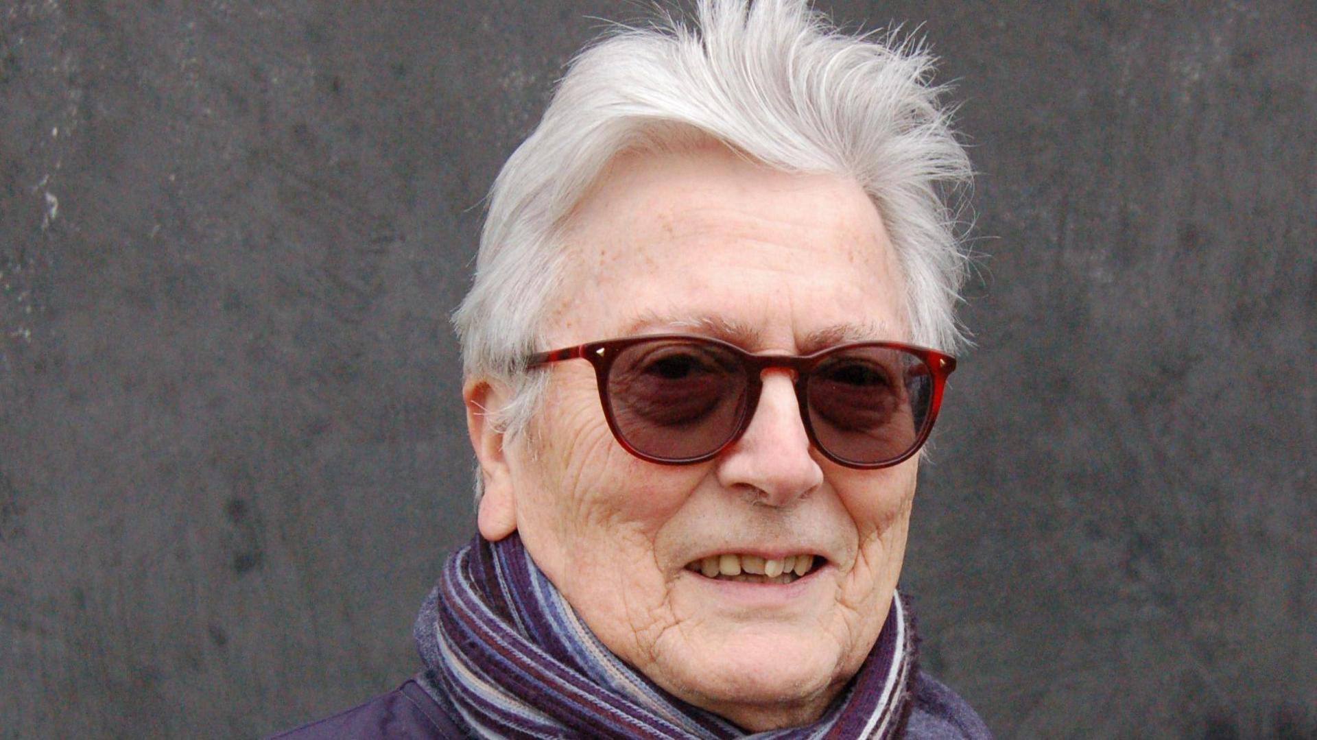 Richard Fletcher. He has short grey hair and is smiling at the camera. He has red sunglasses on as well as a blue coat with a blue scarf.