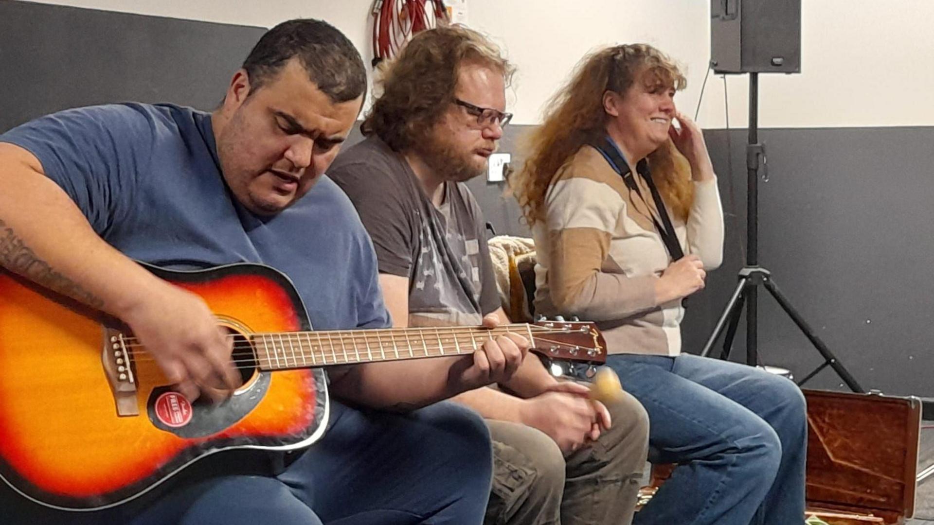 Gaynor sits to the far right of the image, next to two men who are also in the band. Gaynor is smiling and wears a beige striped long sleeve top with blue flare jeans. She sits on a bench next to a man with mid length red hair and wearing black wire framed glasses. He looks down as he plays a maraca and wears a pale brown short sleeve t shirt with khaki green combat trousers. Next to him is a man with black short shaved hair, wearing a blue short sleeved t shirt and jeans, he is playing a guitar.