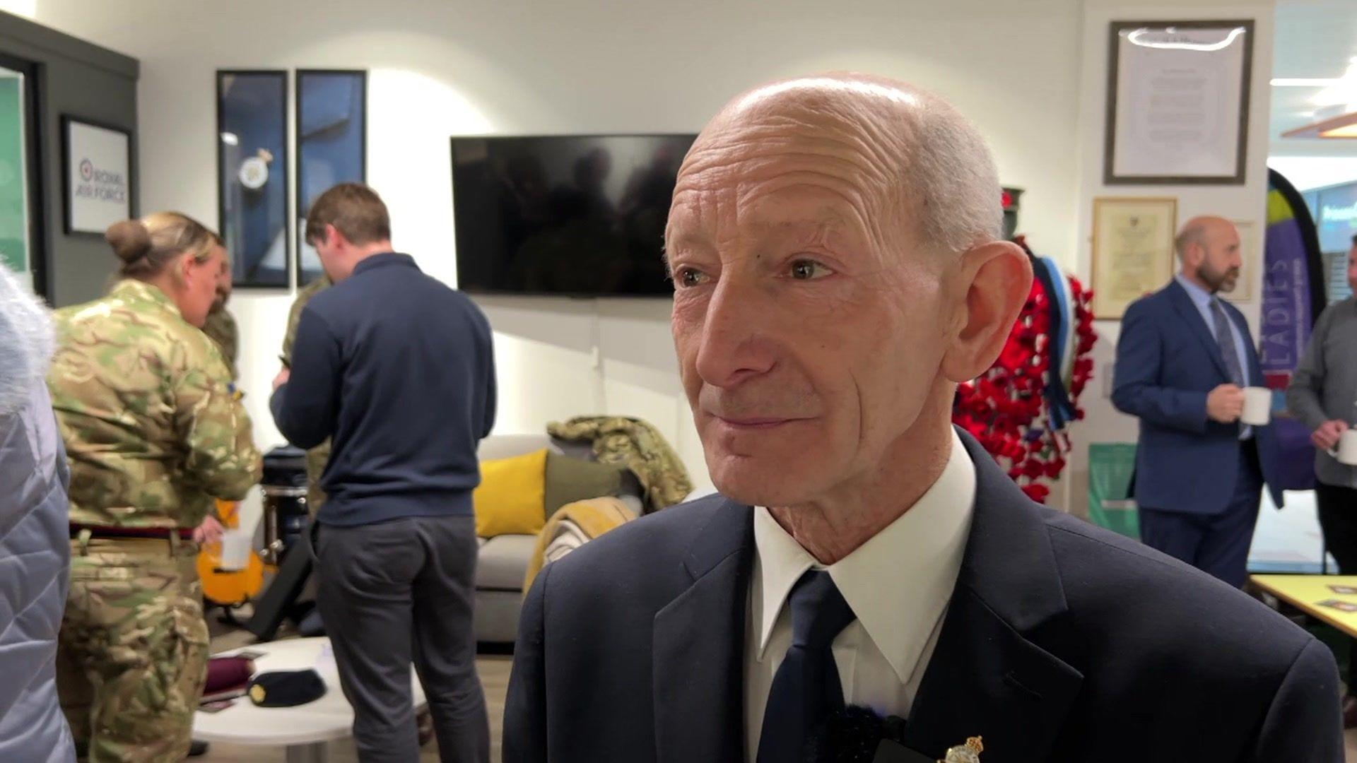 Mr Johnson wears a navy blue suit and a navy pin on his lapel. He is mainly bald with grey receding hair. He is talking to the camera with other people in the background in conversation.