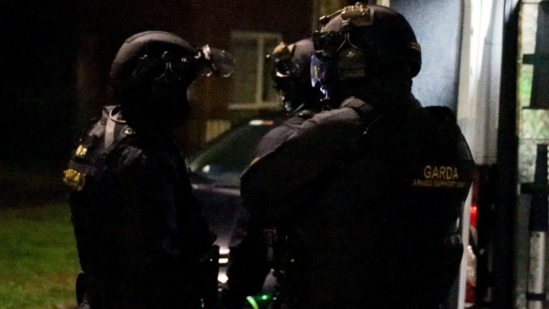 Garda officers outside a building at night - three officers are wearing full black Garda uniforms with a badge that reads 'GARDA ARMED SUPPORT UNIT', they are all wearing black helmets and talking to each other. 