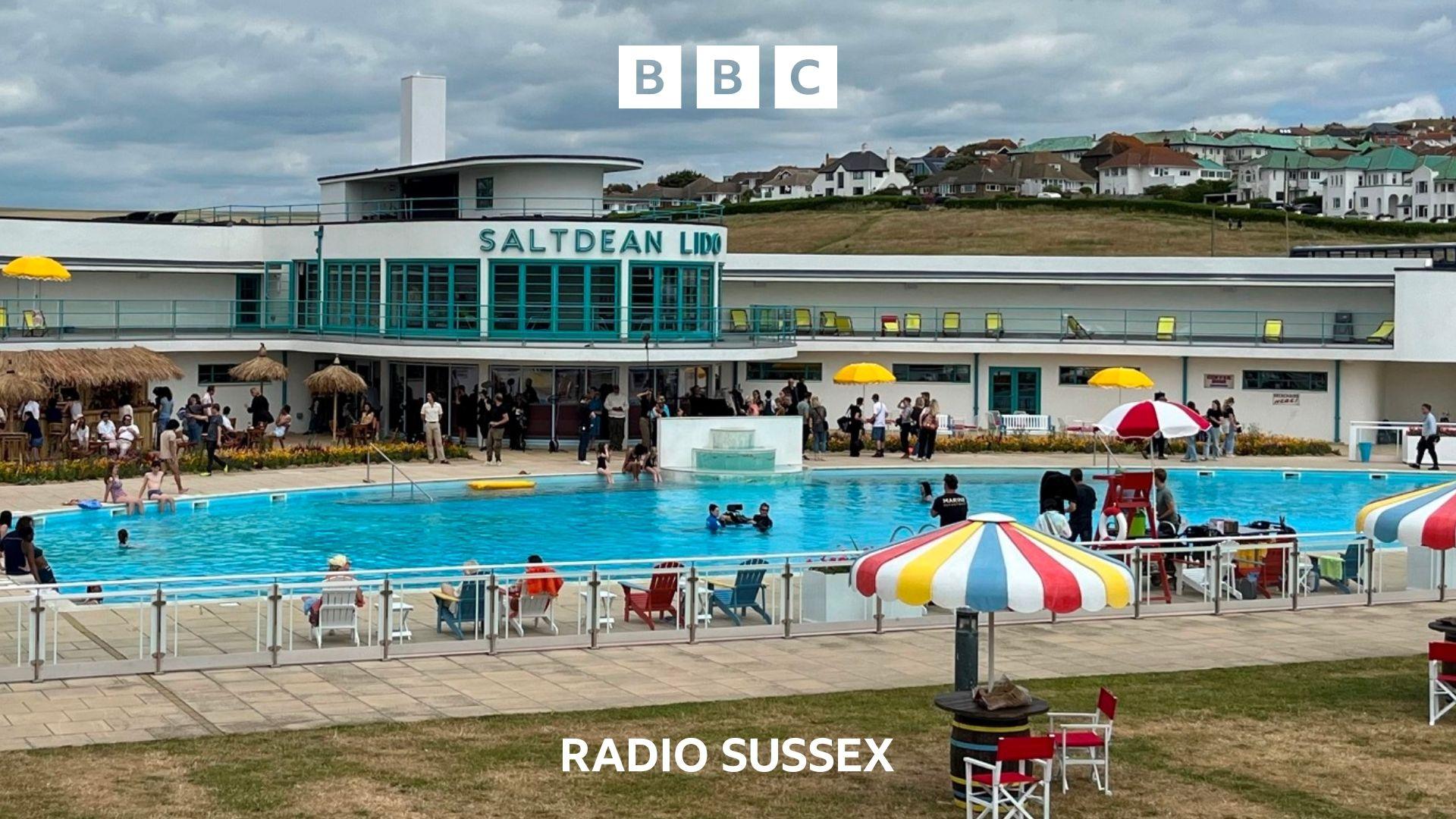 An exterior shot of Saltdean Lido. stripey parasols  and red fold up chairs are dotted around.  Extras and film crew are scattered about.