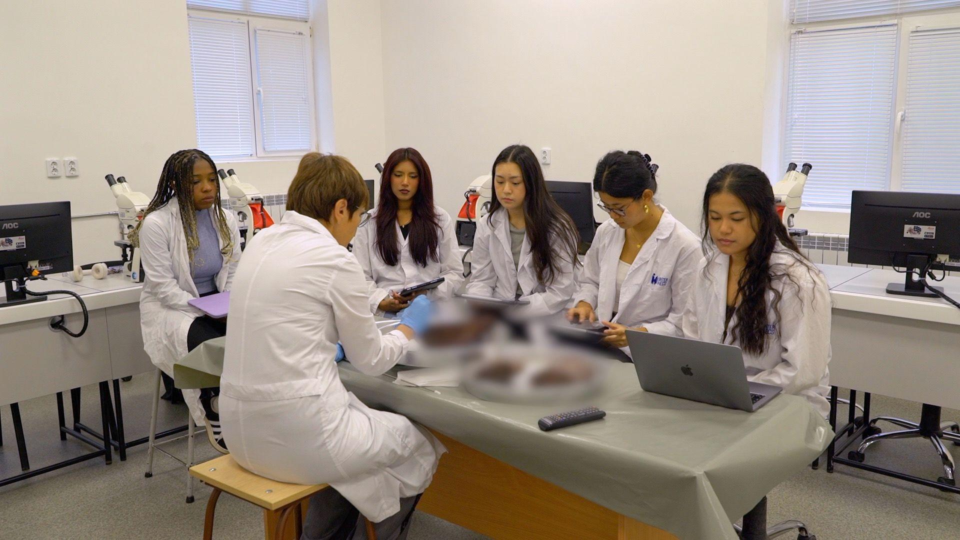 A group of five medical students, wearing white medical scrubs, gather around a desk where their teacher talks them through an examination.