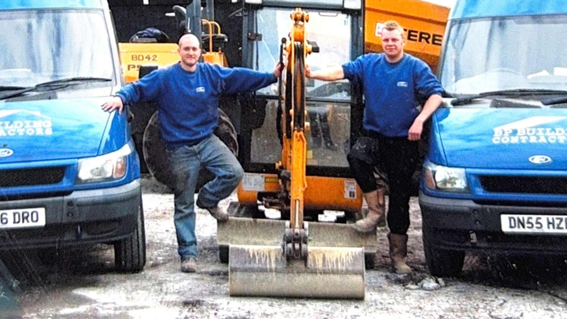 Tow men in matching blue jumpers with a white left breast logo stand either side of a yellow JCB digger and between two identical blue Ford vans. The vans have a white building firm logo printed on the bonnet. The man on the left is bald and is wearing blue jeans and black boots. The man on the right has short light hair, black work trousers and brown boots.