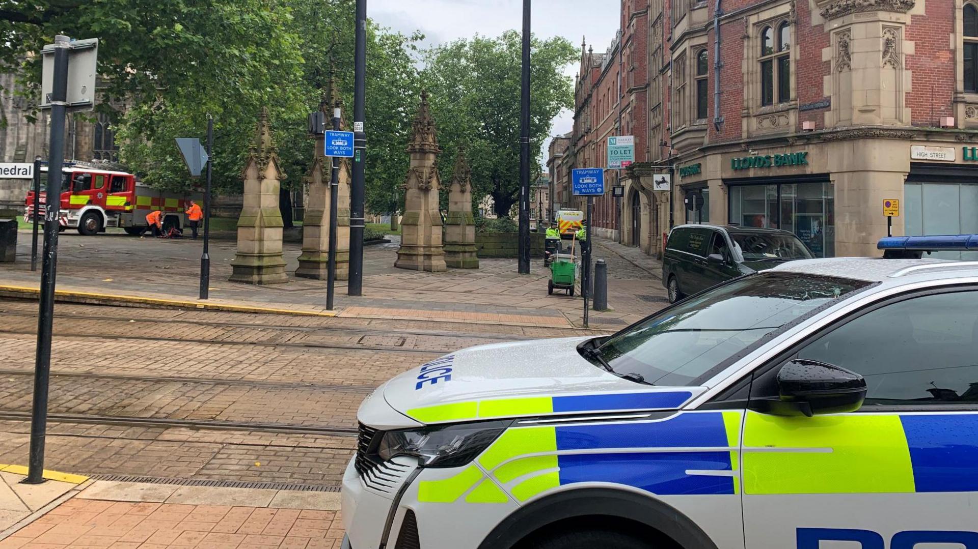 A police car parked next to the tram tracks in Church Street, Sheffield. A fire engine is parked to the right of the cathedral and a police van can be seen in the alley next to Lloyds Bank.