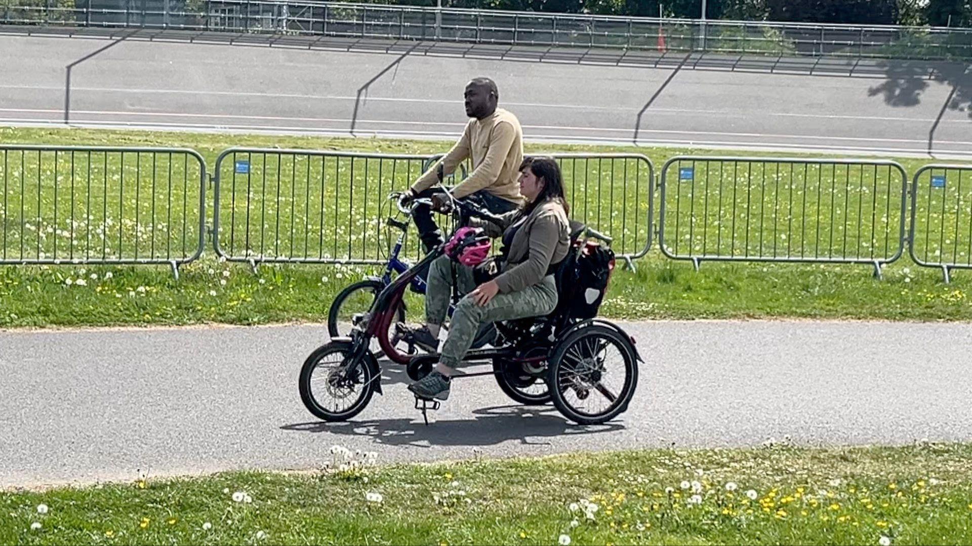 Two individuals ride specially adapted bicycles along a path lined with green grass and safety barriers.