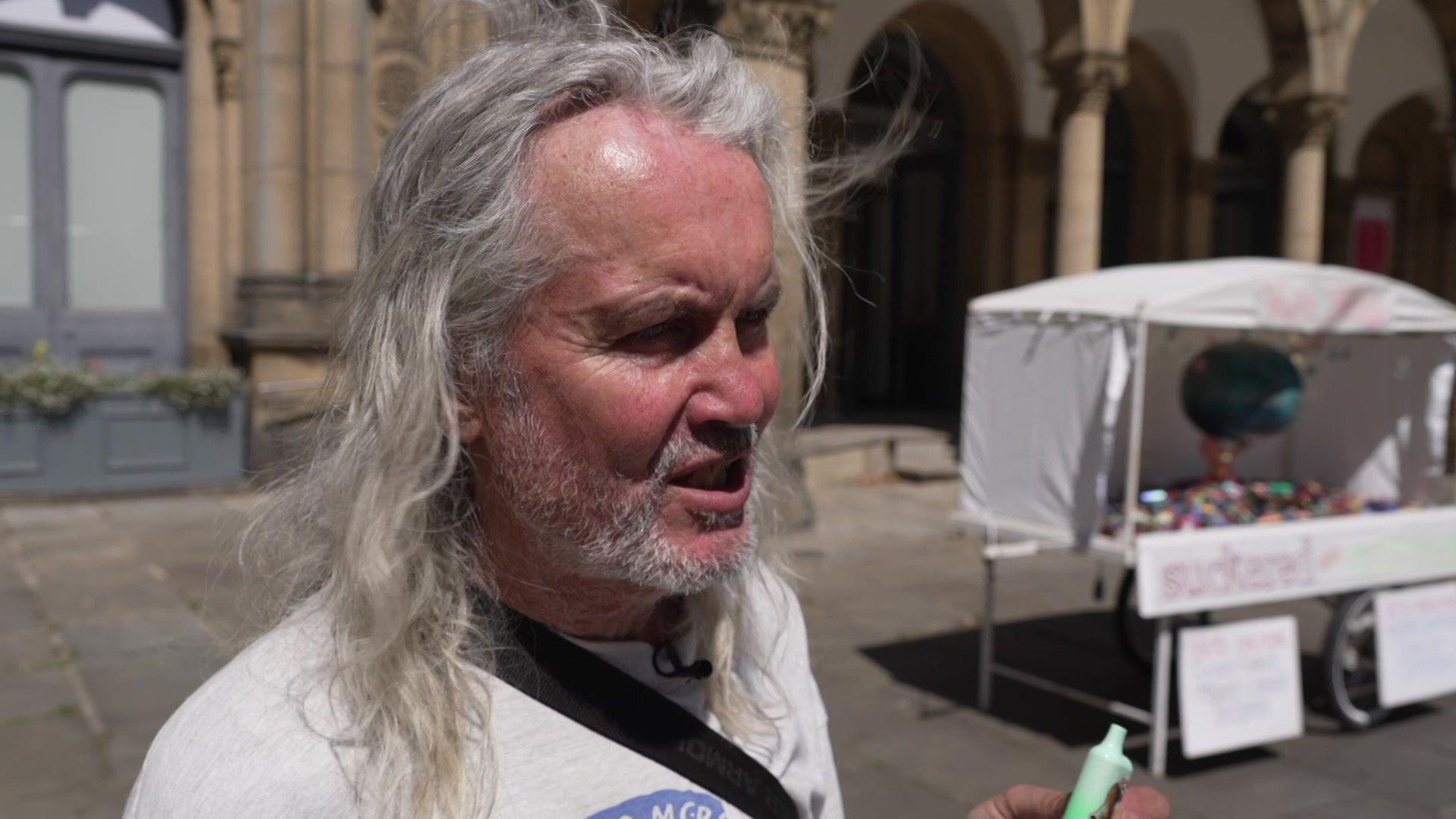 A man with long grey hair holds up a green vape. Behind him is a vape sculpture