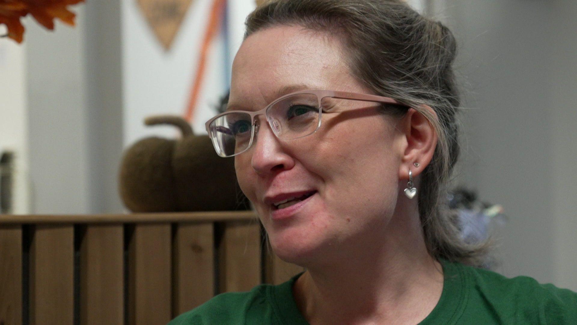 Rebecca Goodier has light brown hair which is tied up and is wearing light pink framed glasses and a green t-shirt and a heart shaped pendant earring.