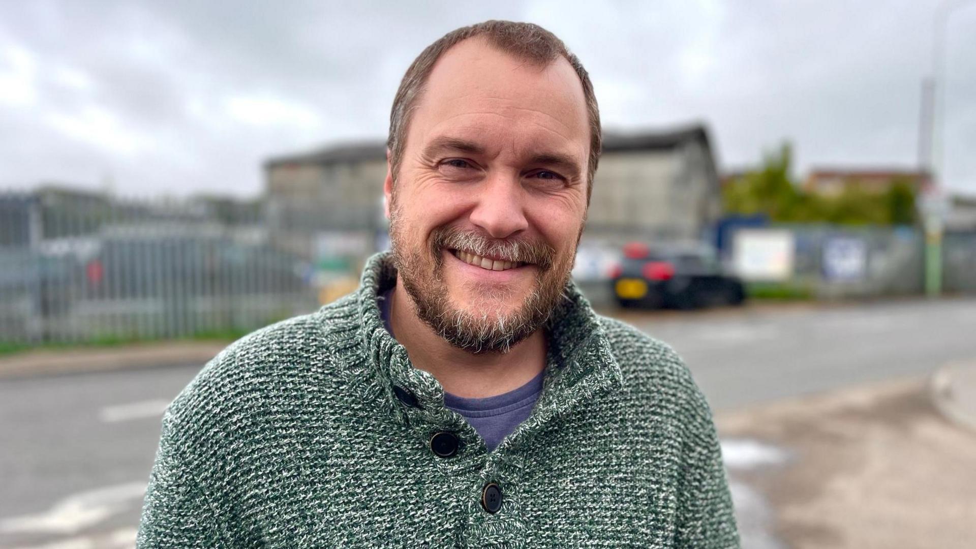 Philip Cook with short light brown hair and a short beard, wearing a white flecked fisherman-style grey jumper, with buttons at the neck and behind him, across a road, a warehouse