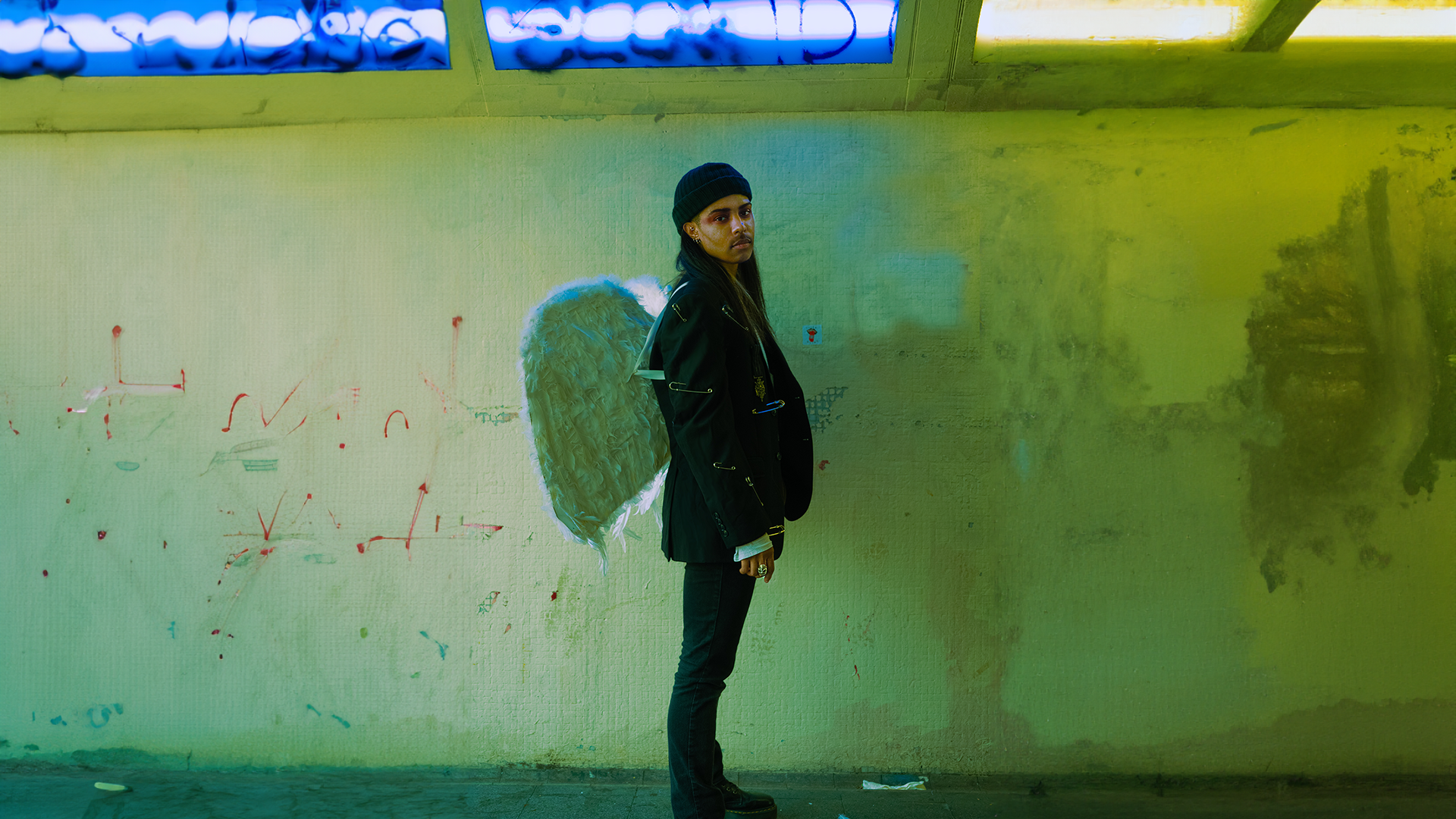 Musician Troi Irons, wearing black and a white pair of wings, standing sideways against a wall