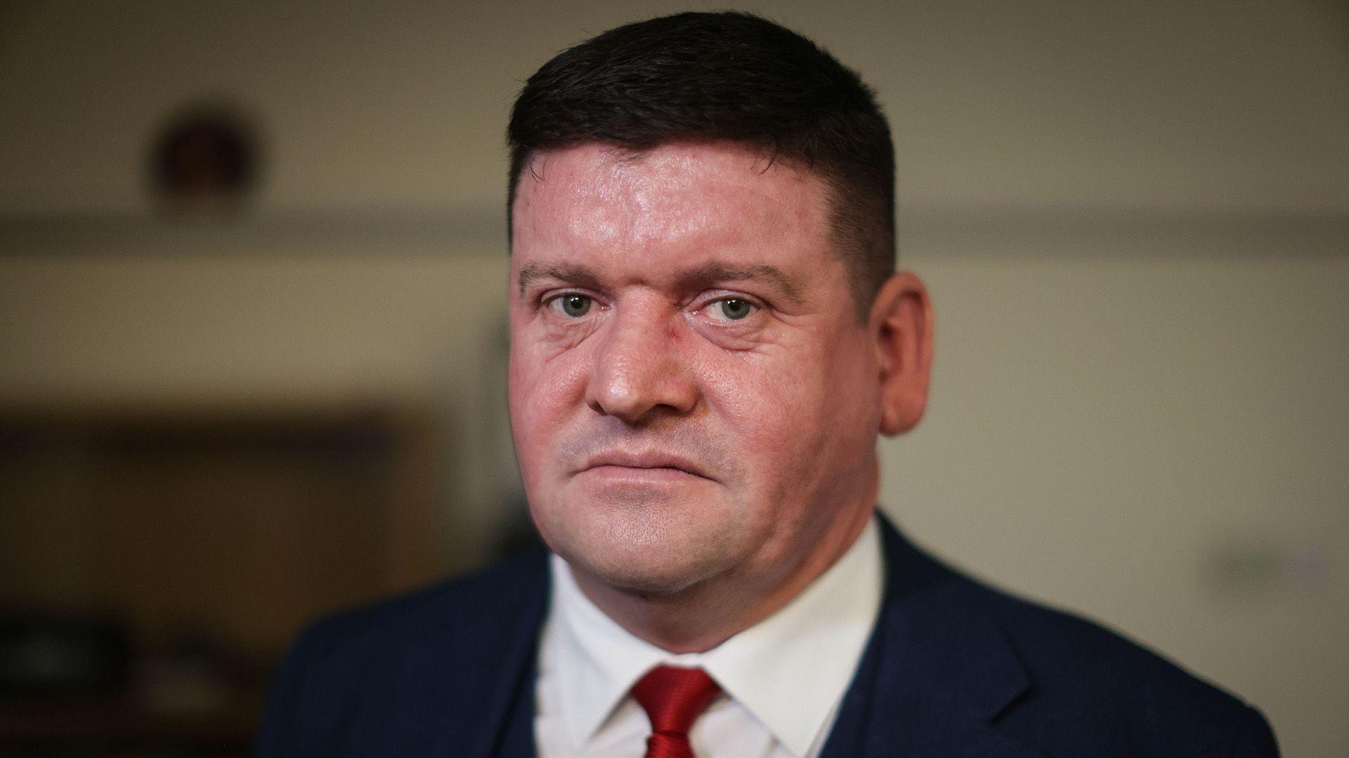 A man in his 40s with short dark hair and wearing a blue suit and red tie