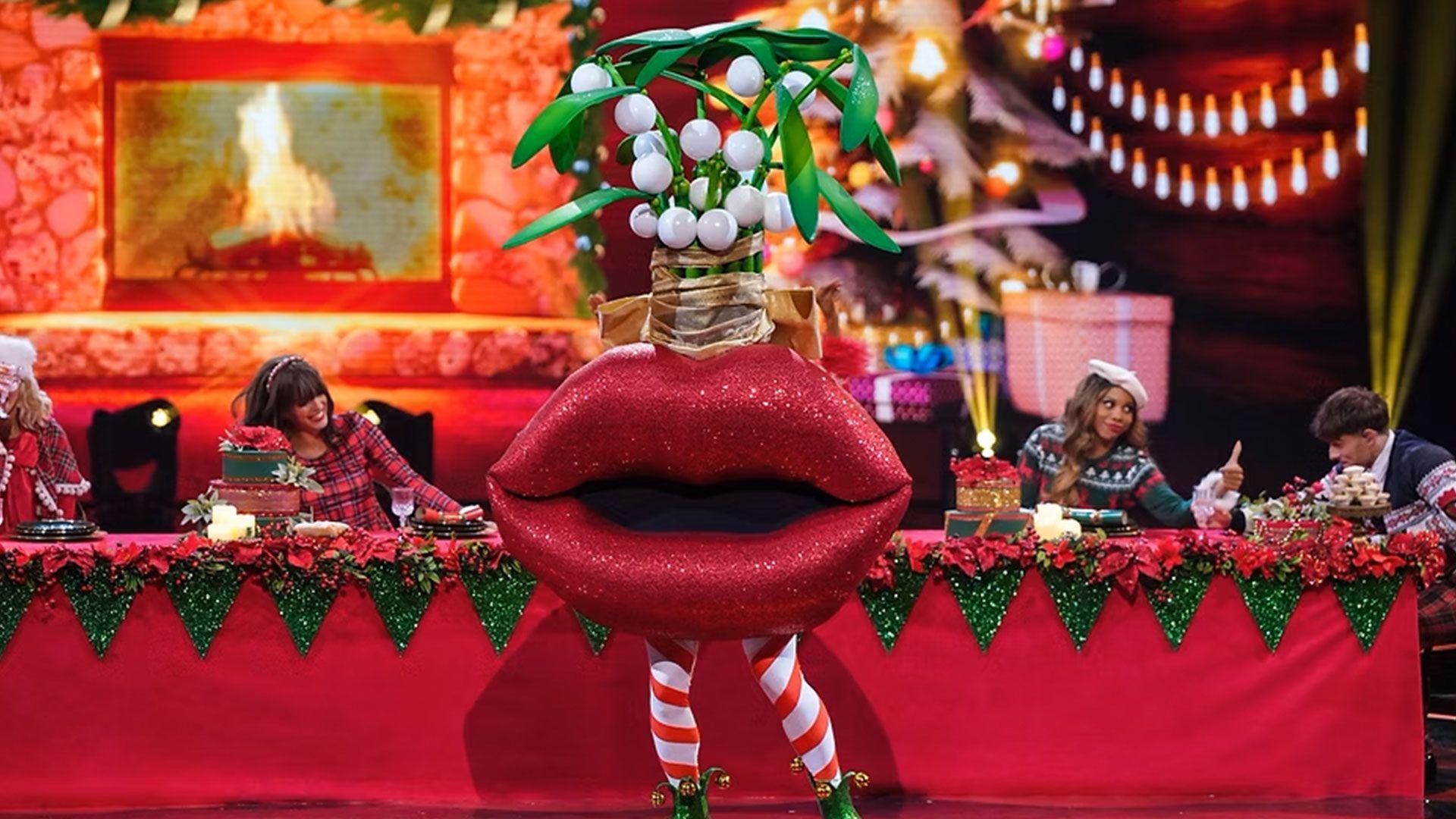 A singer dressed as a large pair of lips with mistletoe dangling over sings next to a Christmas table on a TV set