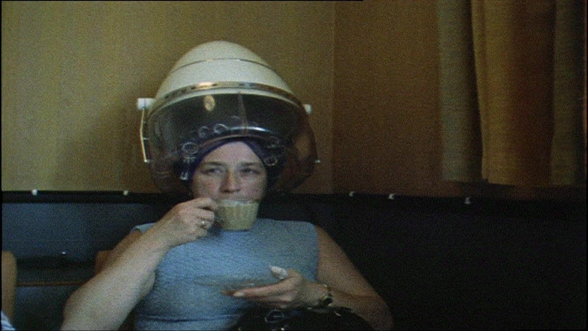 Margaret with rollers in under a standing hair dryer drinking from a cup