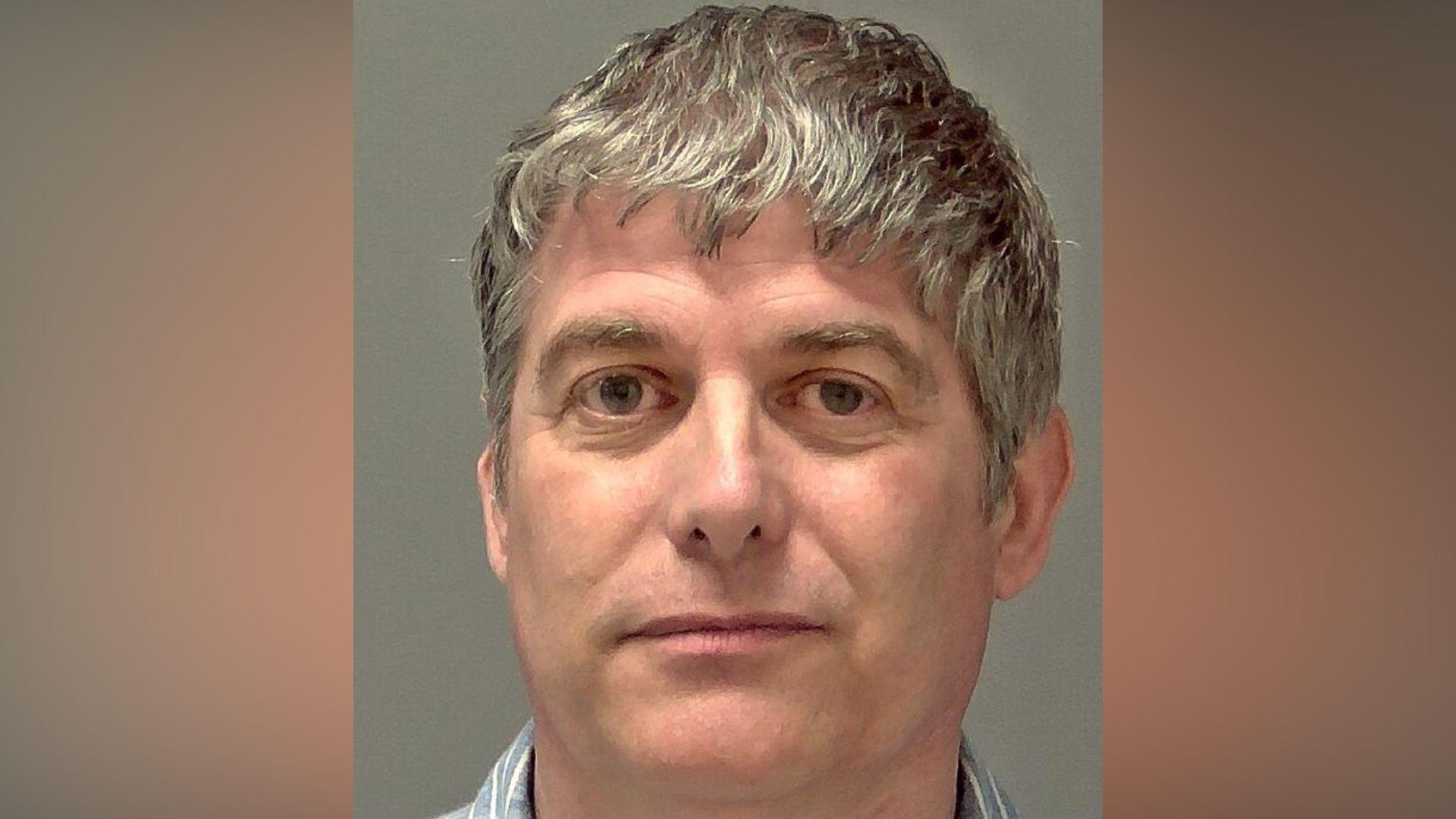 A police custody image of a man who is looking directly at the camera and slightly smiling. He has grey hair that slightly hangs over his forehead and he is wearing a blue shirt with white stripes on it. 