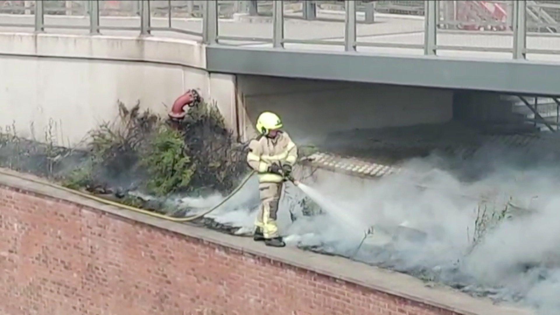 A firefighter, in protective clothing, stands on a wall and sprays water onto a smouldering fire with grey smoke. The grey structure of a bridge is visible in the background.