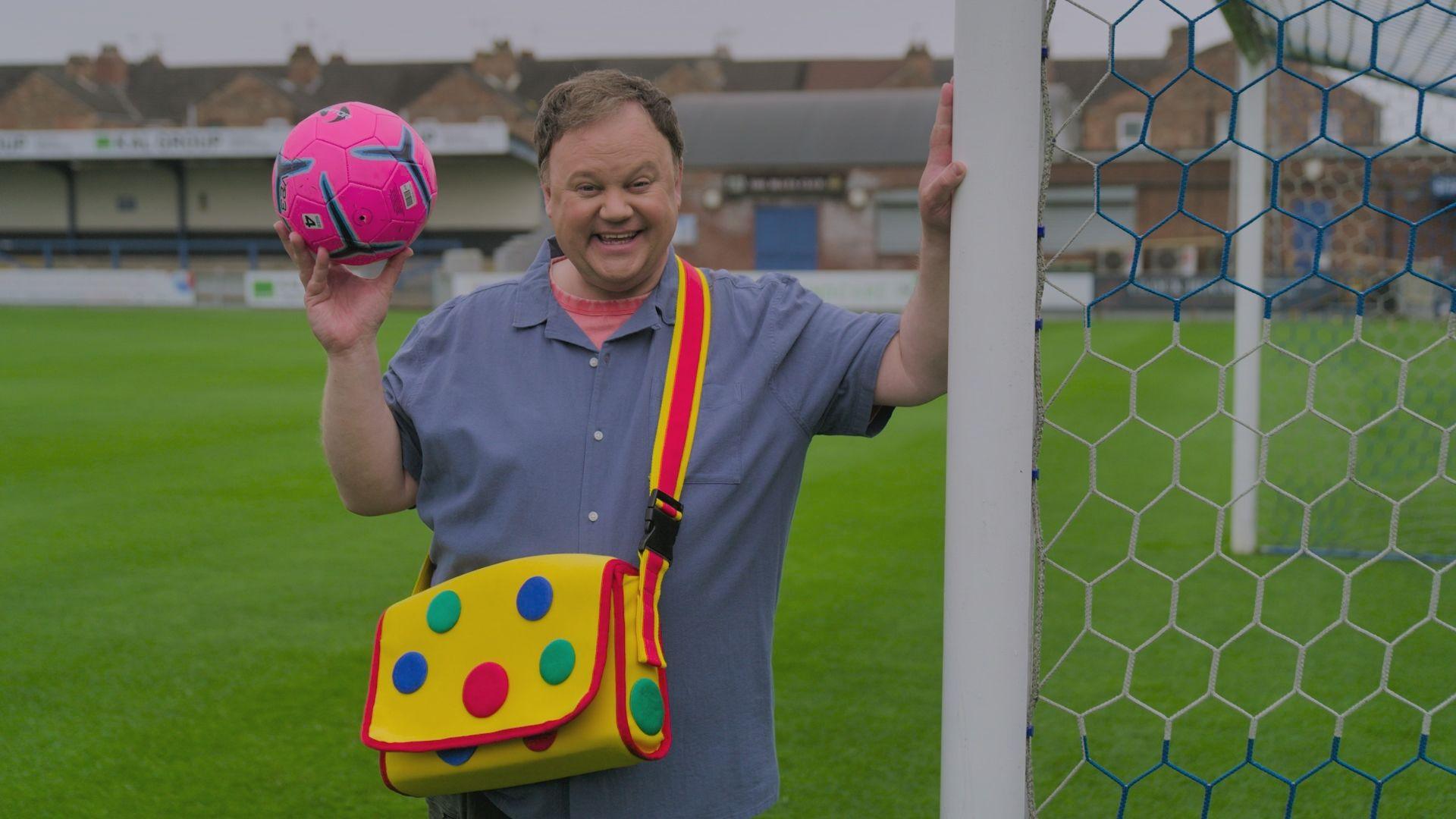 Justin Fletcher on a football pitch holding a pink football and wearing Mr Tumble's spotty bag.