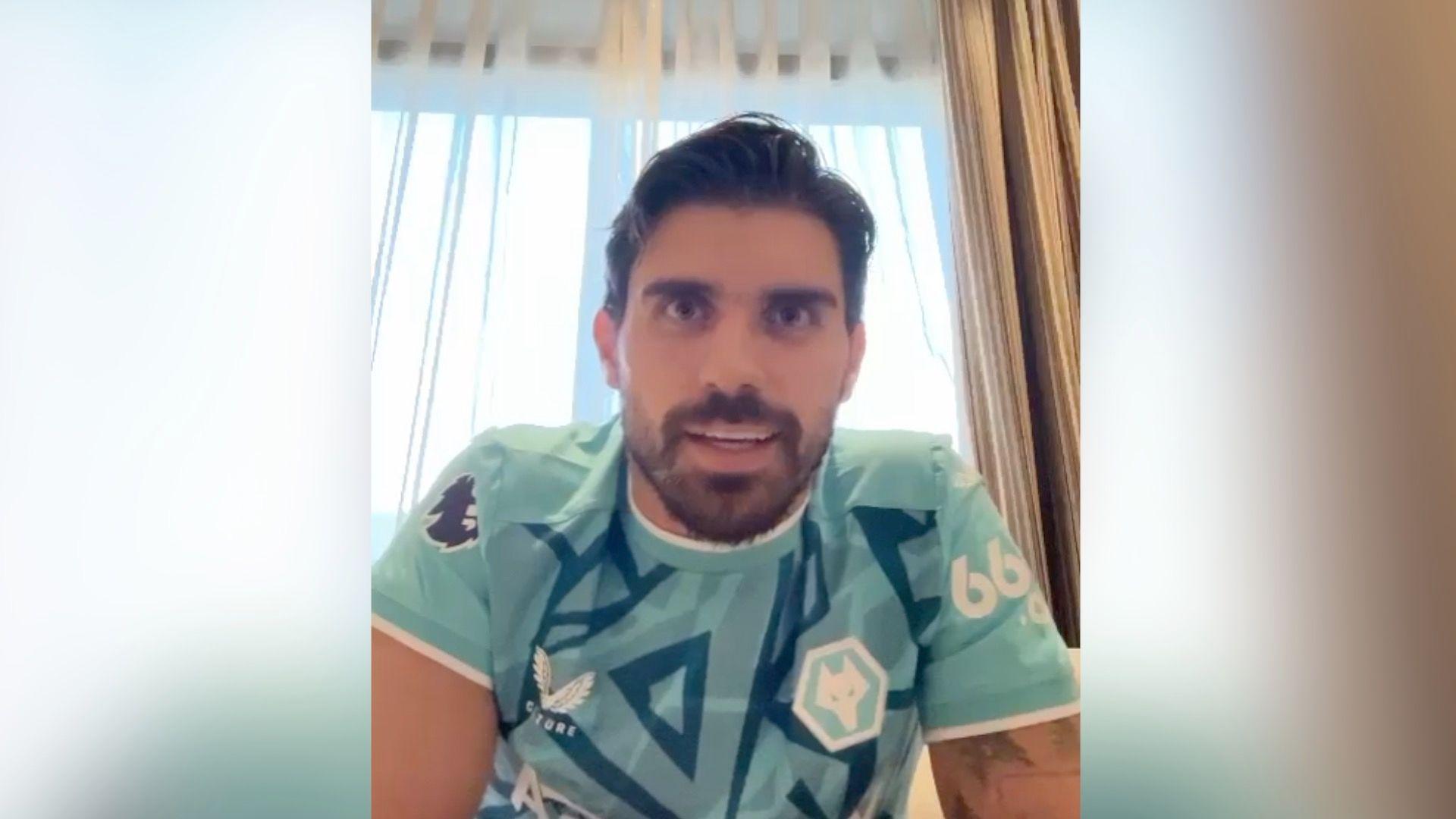 A man with a beard and moustache in a Wolves football shirt sat in his living room. The shirt is green with blue diamonds and lines.