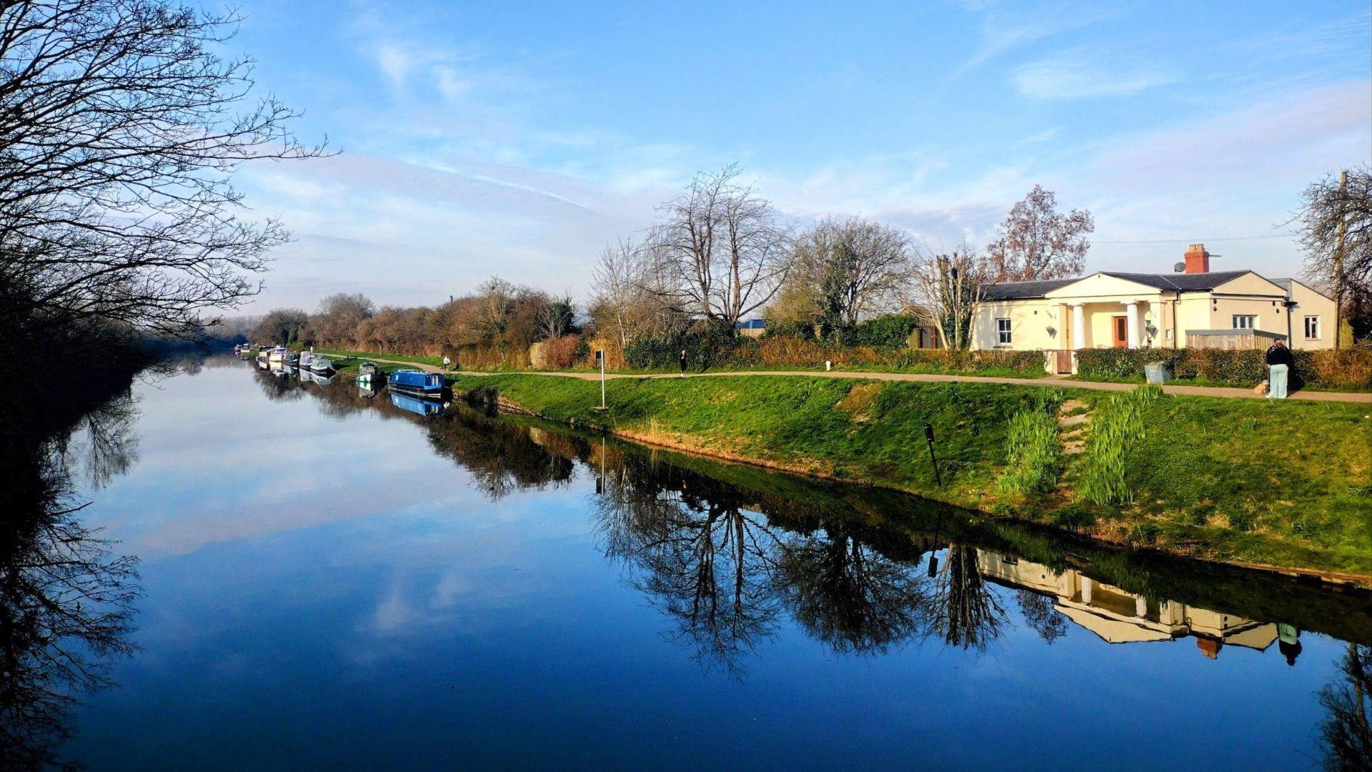 A blue sky with a blue reflection in the water on a canal in Gloucestershire. The water is still and a cream-coloured house is seen on the bank