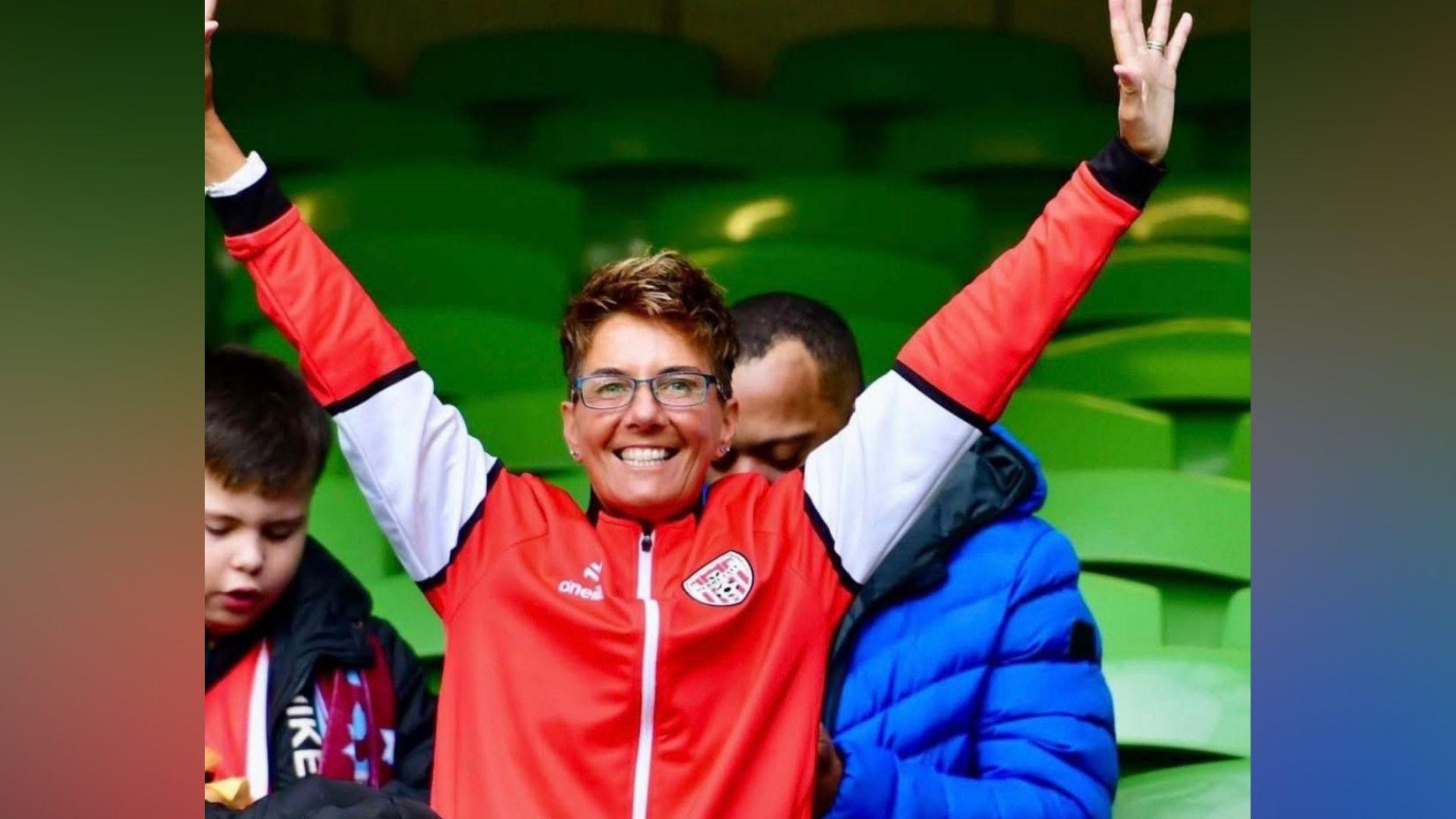 A woman smiling with her hands int he air wearing a red and white zipped up jacket. She has glasses and short brown hair. 
