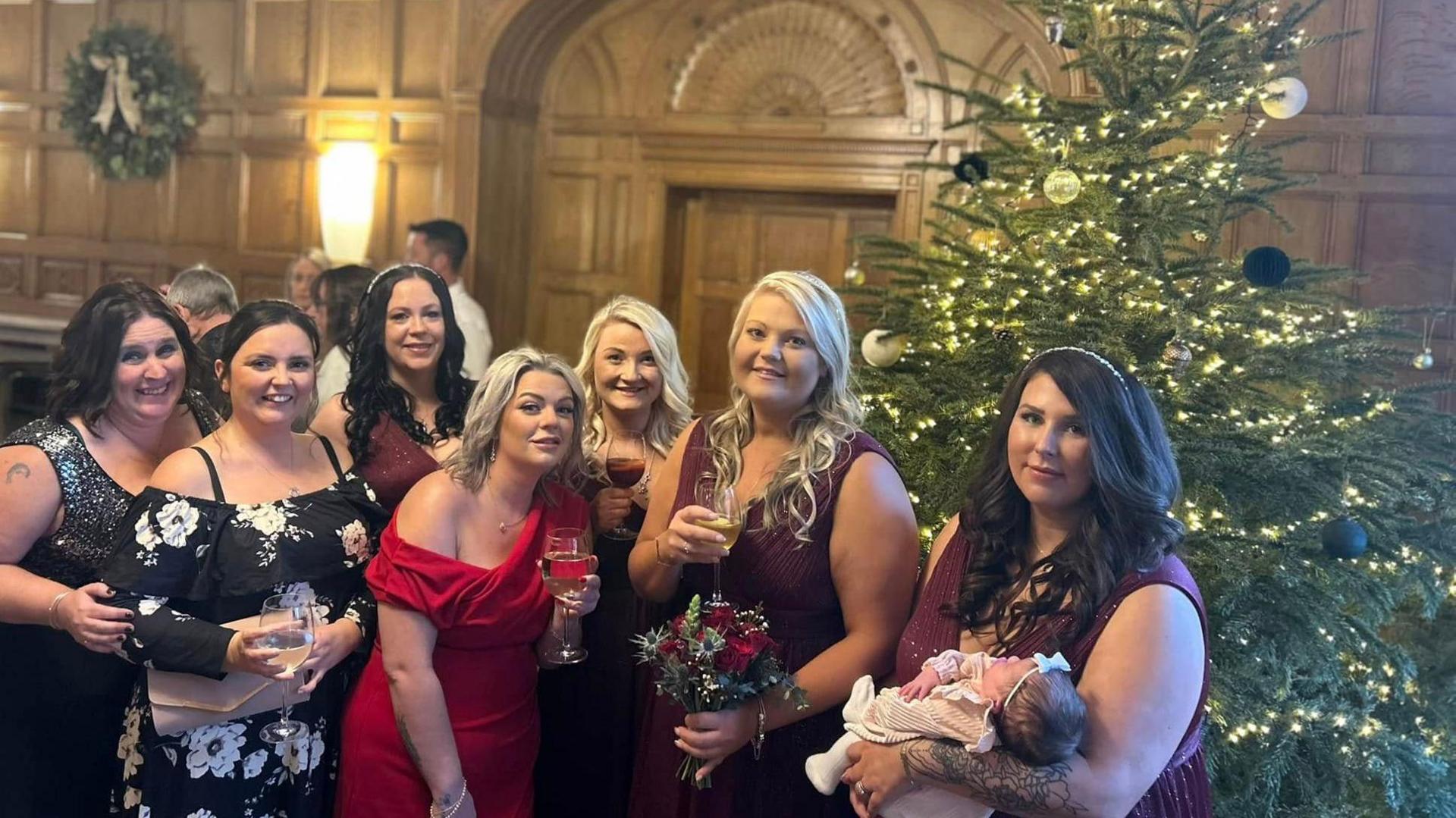 Seven women and a baby pose in front of the Christmas tree set up in the wood-panelled venue where the Hollidays had their wedding