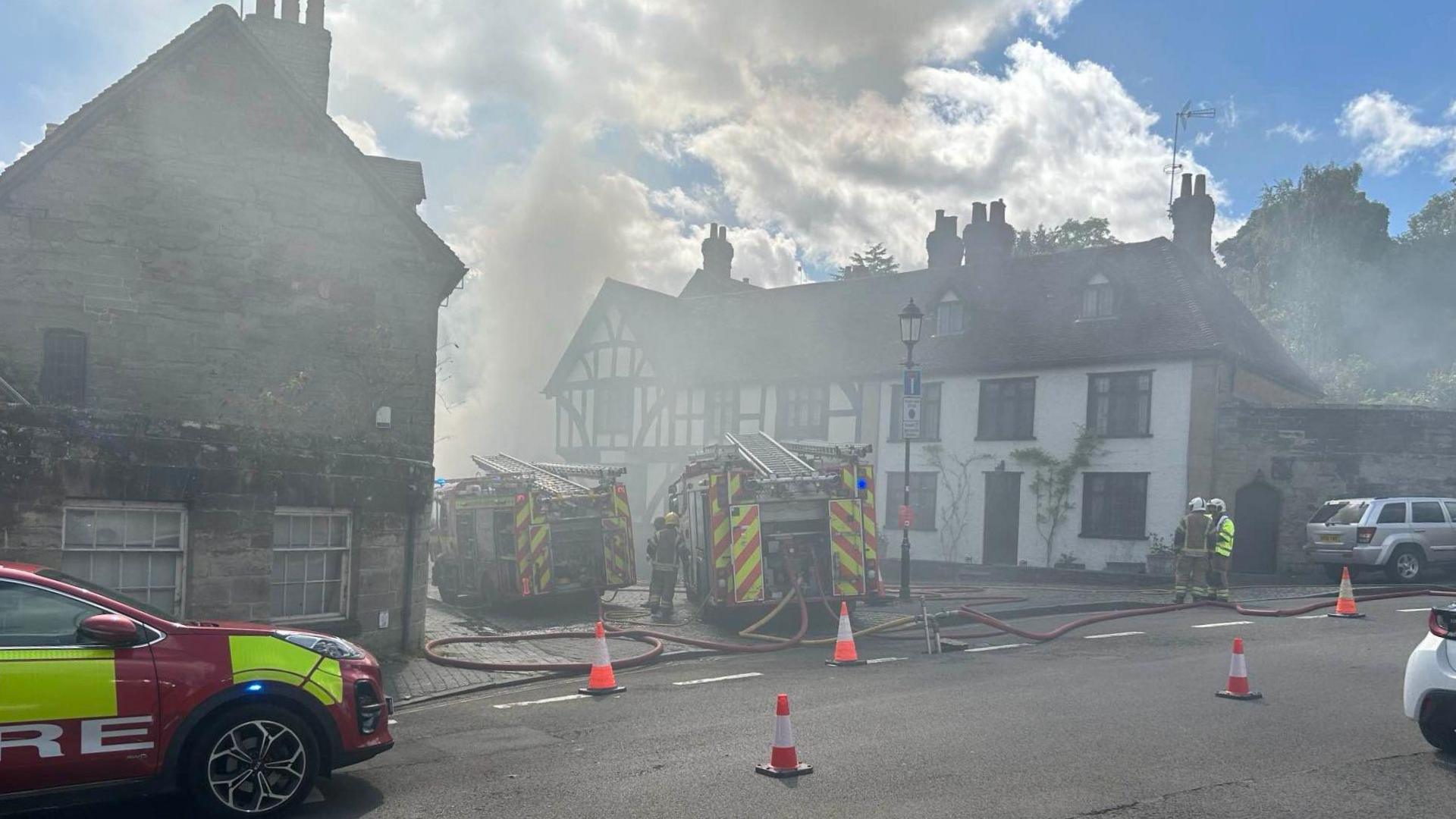 Grey smoke rises into the sky from wooden buildings. In the road there are red traffic cones. There is a red and neon yellow fire car, and two fire engines parked near the smoke, with hoses trailing on the floor