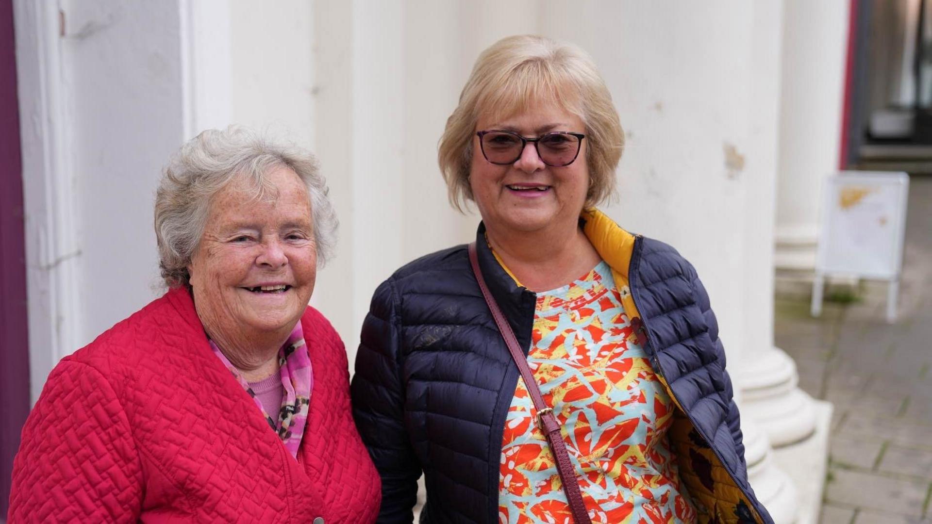 Two women one in a red coat, one in a blue coat, smile for the camera. 