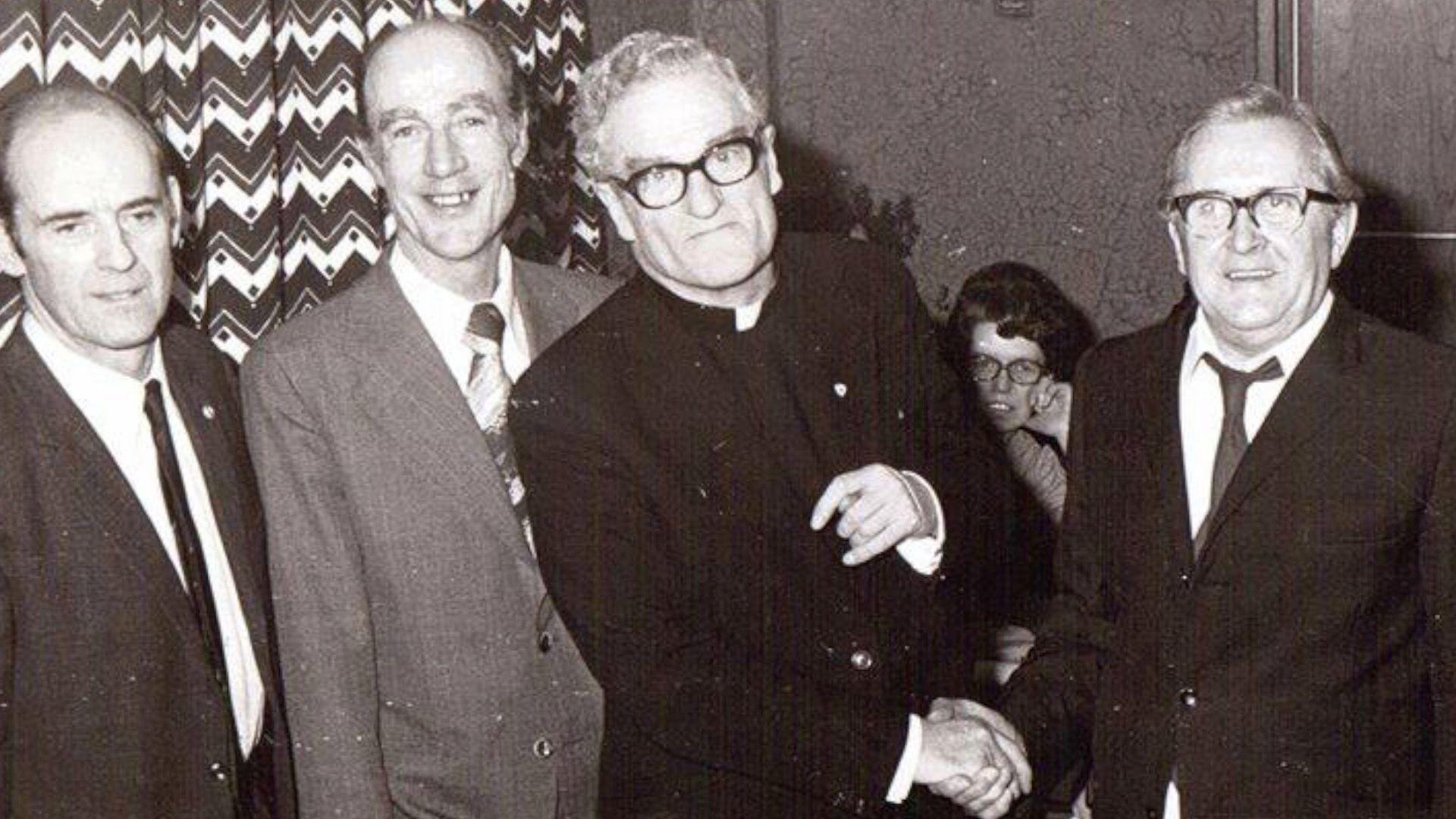 Fr Austin Eustace in a black and white photo with three other man and a woman in the back ground. Fr Austin is wearing his black suit with priest collar. He has white/grey curly short hair and is wearing black-framed glasses. He is shaking the hand of a man to his left but looking directly at the camera. 