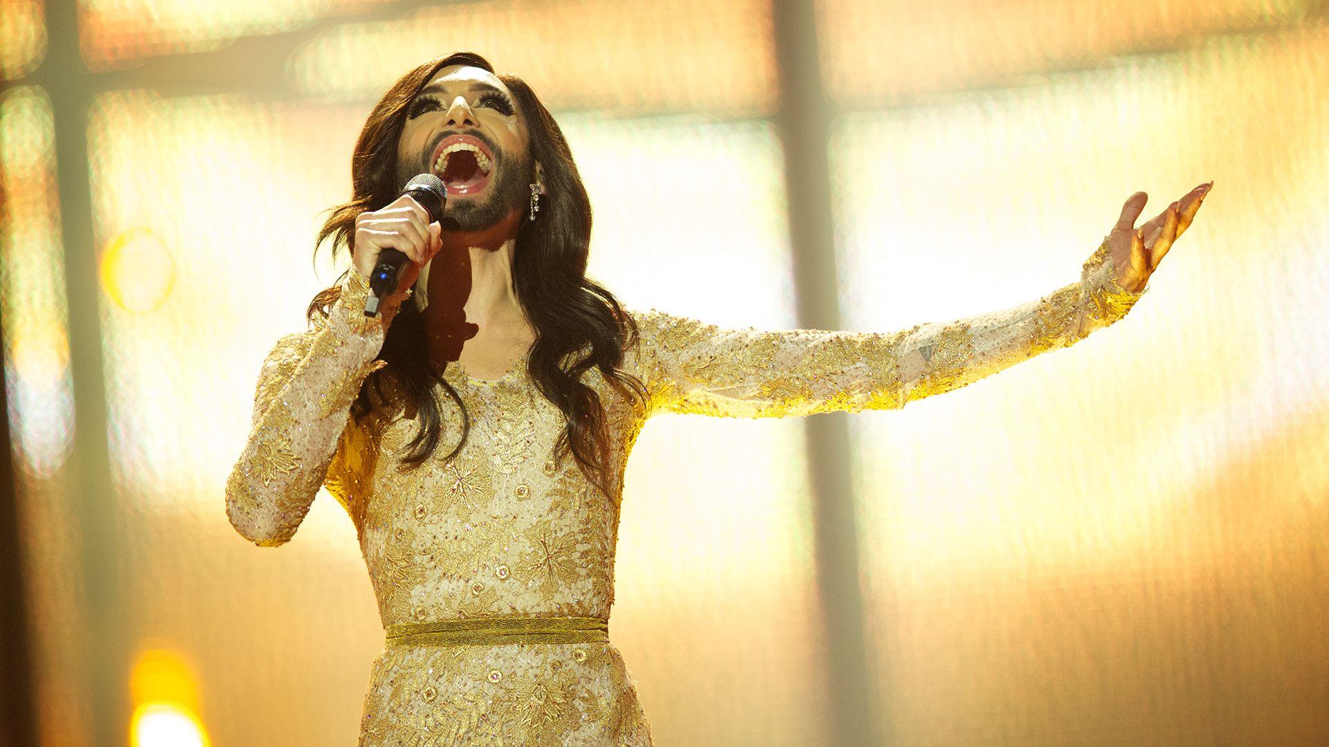 bearded drag queen conchita wurst performing at the 2014 eurovision song contest. she is wearing a gold brocade dress, has long dark hair and is holding a microphone while looking upward