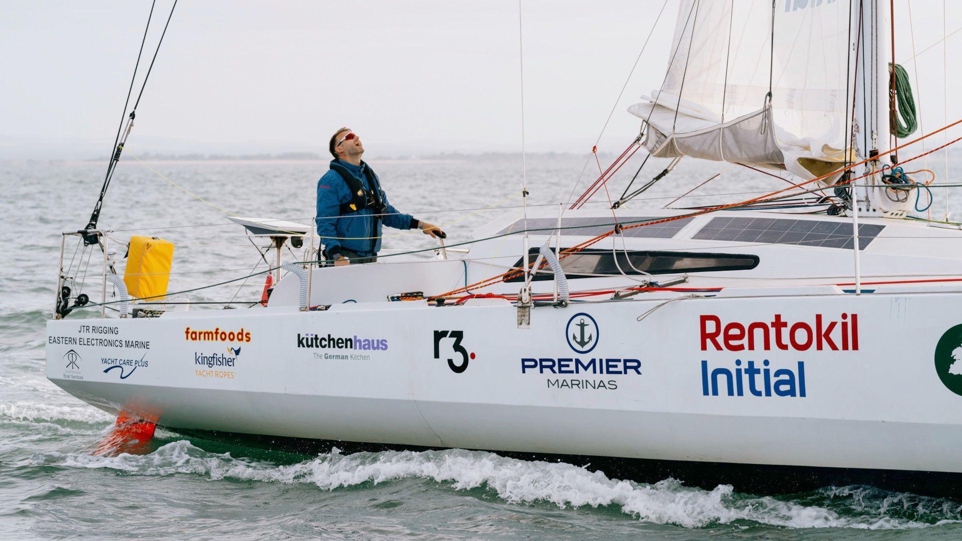 Mr Ketchell is sailing aboard Mindset. He is wearing the same outfit, a blue waterproof long sleeved jacket with a black buoyant vest on. He is in the middle of the ocean and his yacht is while with various business sponsor logos on the exterior.